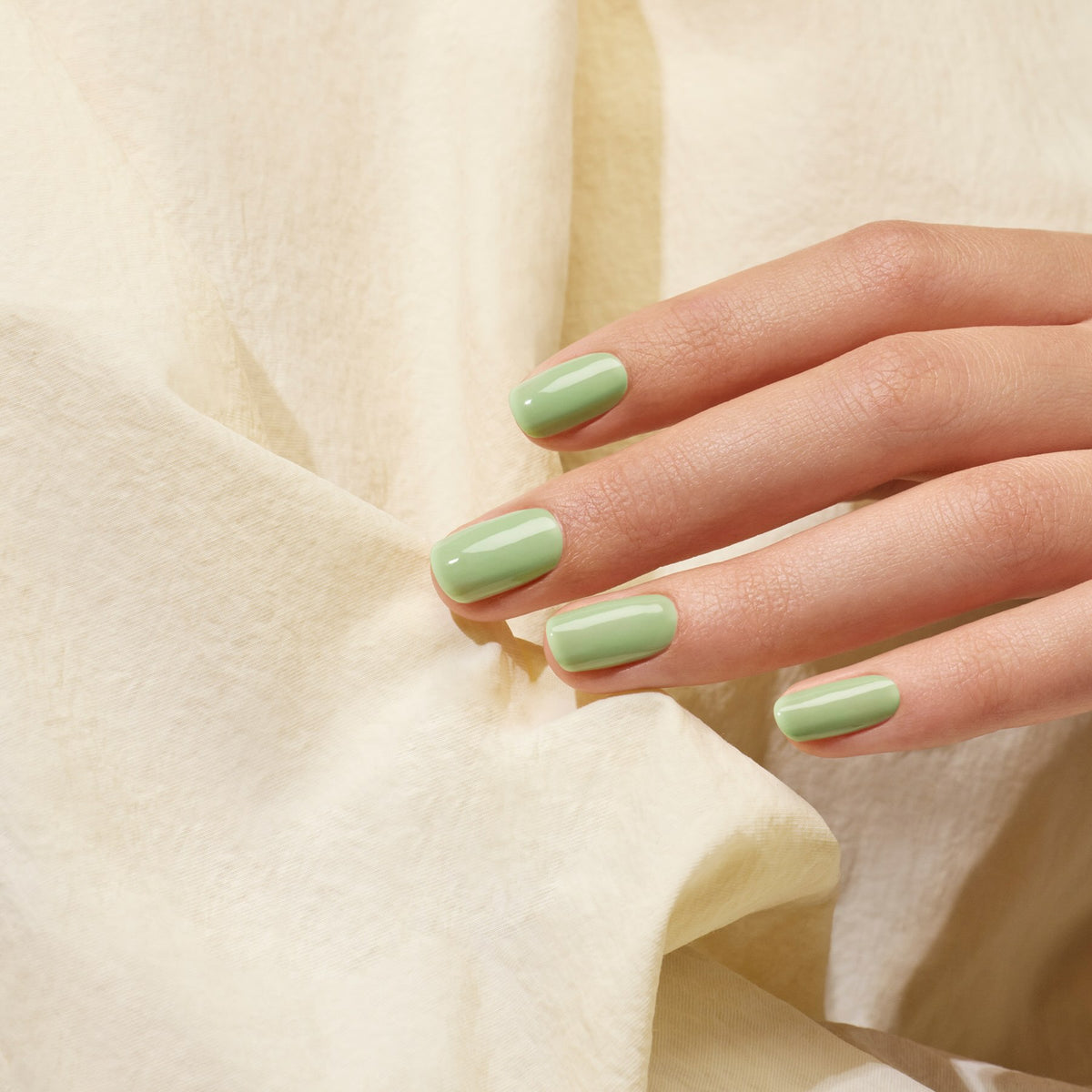A hand with neatly manicured nails painted in Manucurist’s Milky Matcha gel polish rests gently on a cream-colored, textured fabric background, evoking creamy nuances and subtle botanical notes.