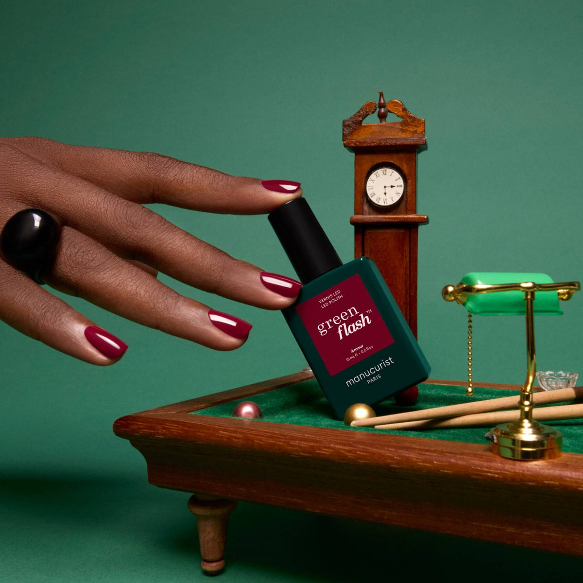 A hand with red polished nails holds a bottle of Manucurist Green Flash gel polish in the shade Amour near a small pool table, clock, and balance scale, all set against a green background.