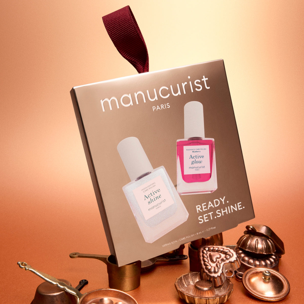 Une boîte du duo format voyage Active™ de Manucurist, contenant deux flacons étiquetés Active Shine et Active Glow, est posée parmi de petits objets en laiton sur une surface brune, devant un fond en dégradé doré et pêche. Un cadeau parfait pour les passionnés de soin des ongles.