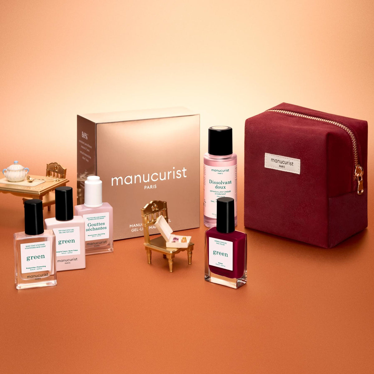 The Manucurist Green™ Discovery Kit Gift Set, featuring naturally derived nail polishes, remover, and a red pouch, is displayed with miniature furniture on a peach background. A gold Manucurist box sits in the back, making this set an ideal holiday gift.