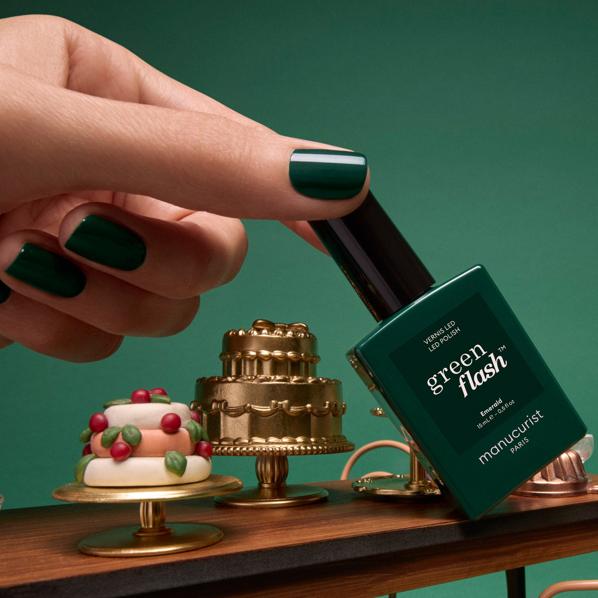 A hand with deep forest green manicured nails holds a bottle of Emerald gel nail polish next to miniature decorative cakes.