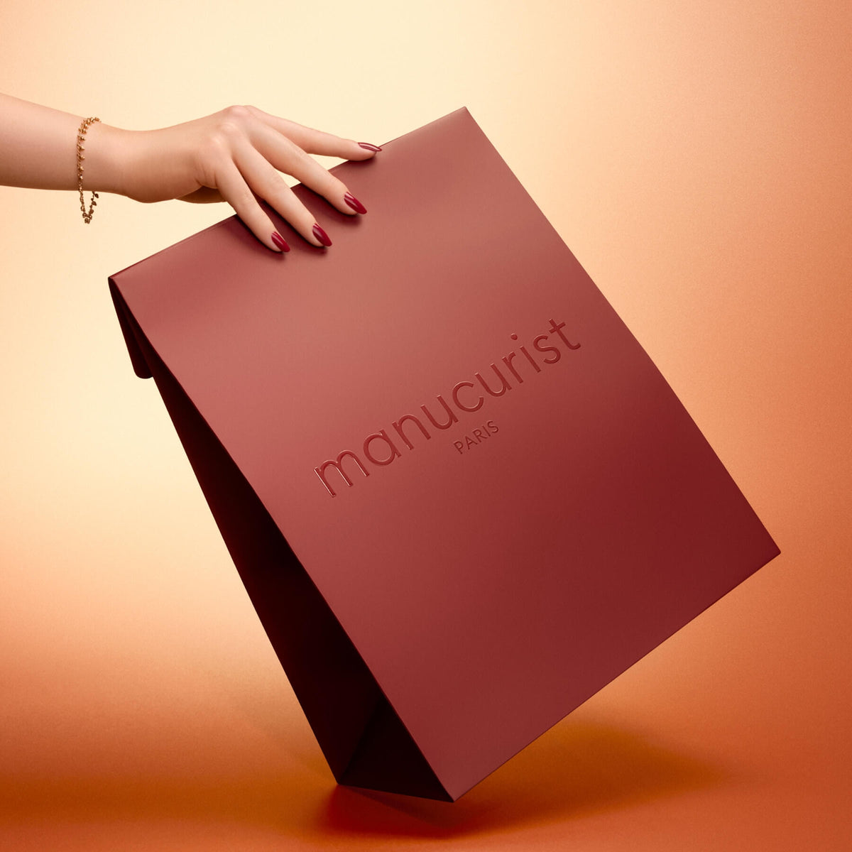 Une main aux ongles vernis portant un bracelet tient le grand Holiday Gift Bag - M de Manucurist, au fini bordeaux mat, sur un fond dégradé pêche.