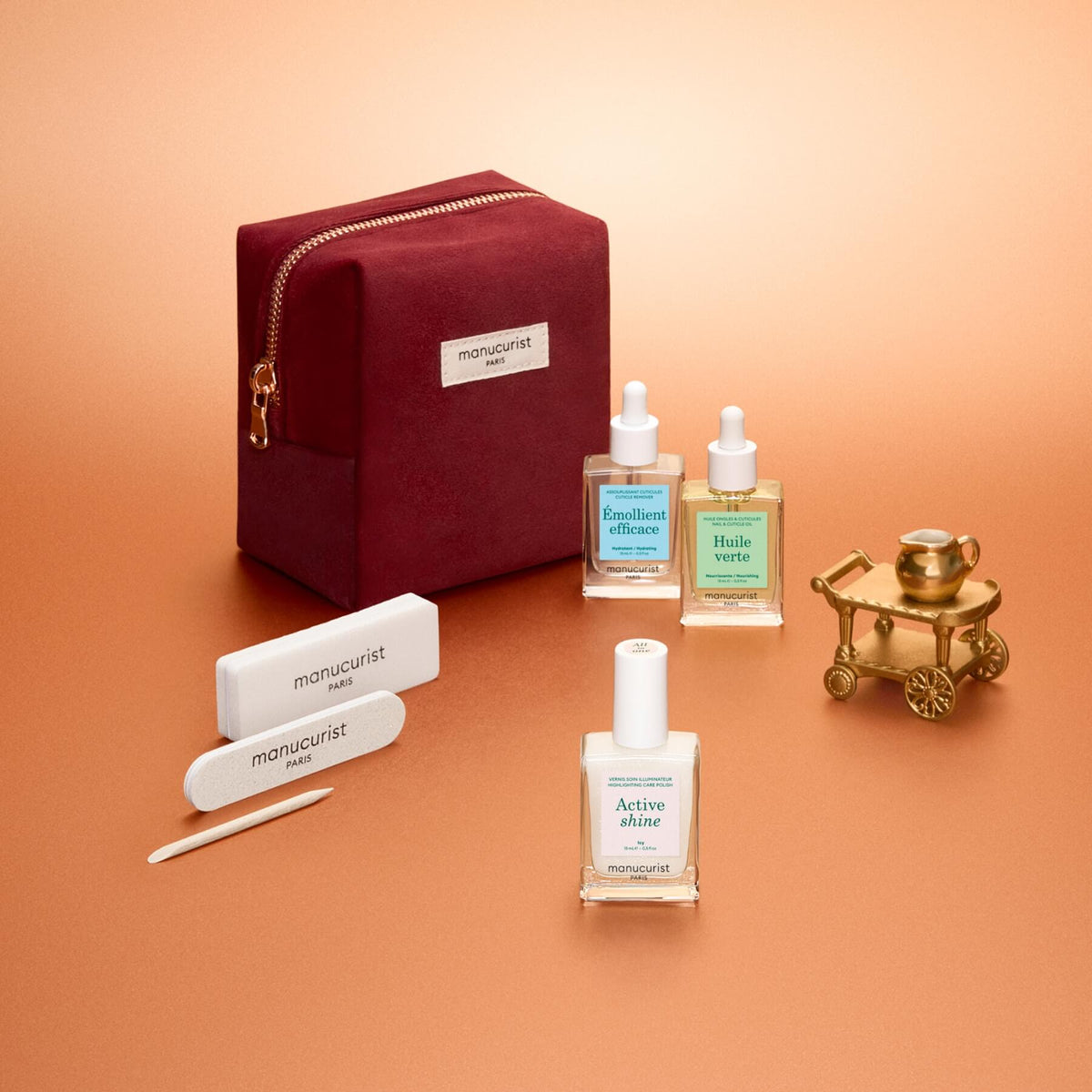The Manucurist Active™ Discovery Gift Set features a burgundy pouch, three bottles of manicure products, a nail file, a cuticle stick, and a small gold cart—all beautifully arranged on a soft orange background to provide everything you need for the perfect manicure routine and stunning nails.