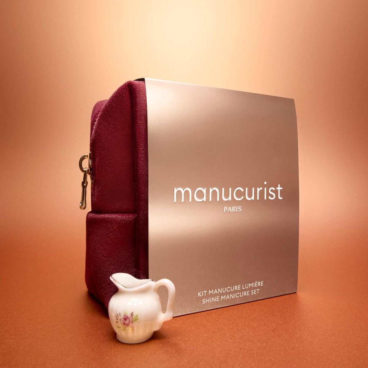 An Active™ Gift Set Holiday by Manucurist, featuring a maroon pouch and a metallic box labeled “Manucurist Paris,” is displayed on an orange surface, accompanied by a small white porcelain creamer decorated with flowers in front.