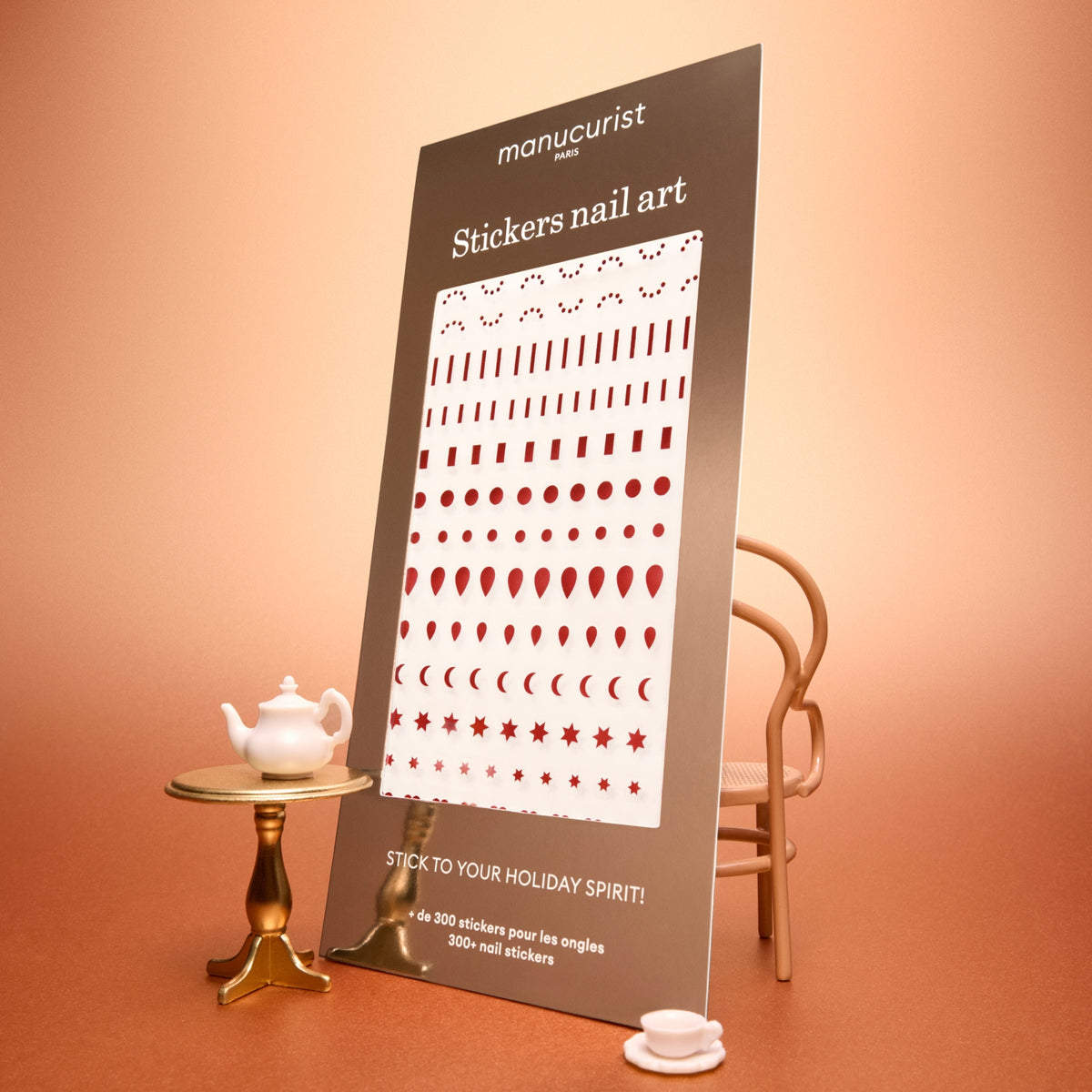 A display board featuring Manucurist Stickers Nail Art Holidays, showcasing festive red shapes on white sheets, is perfect for creating holiday-inspired manicures. The scene is set on a peach-colored surface with a small table holding a teapot and cup, and a chair in the background.