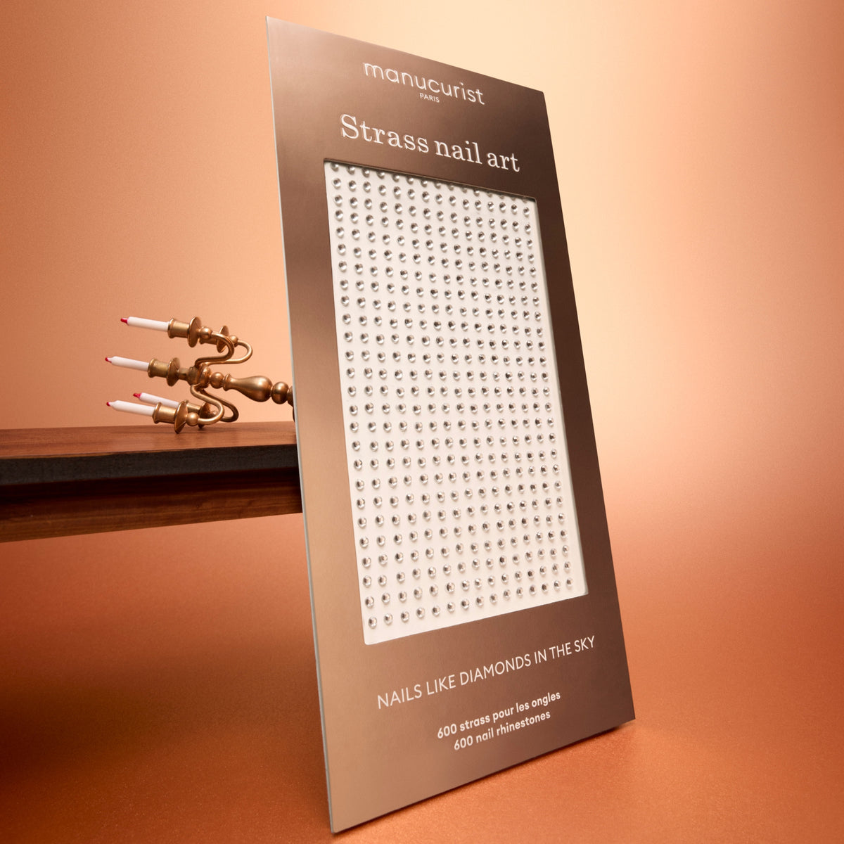 A package of Manucurist Rhinestones Nail Art Holidays is displayed upright on a brown surface, with a modern gold candle holder in the background. Ideal for elevating your next manicure or adding festive holiday flair, the package showcases silver rhinestones arranged in neat rows, reminiscent of professional nail art rhinestone sheets.