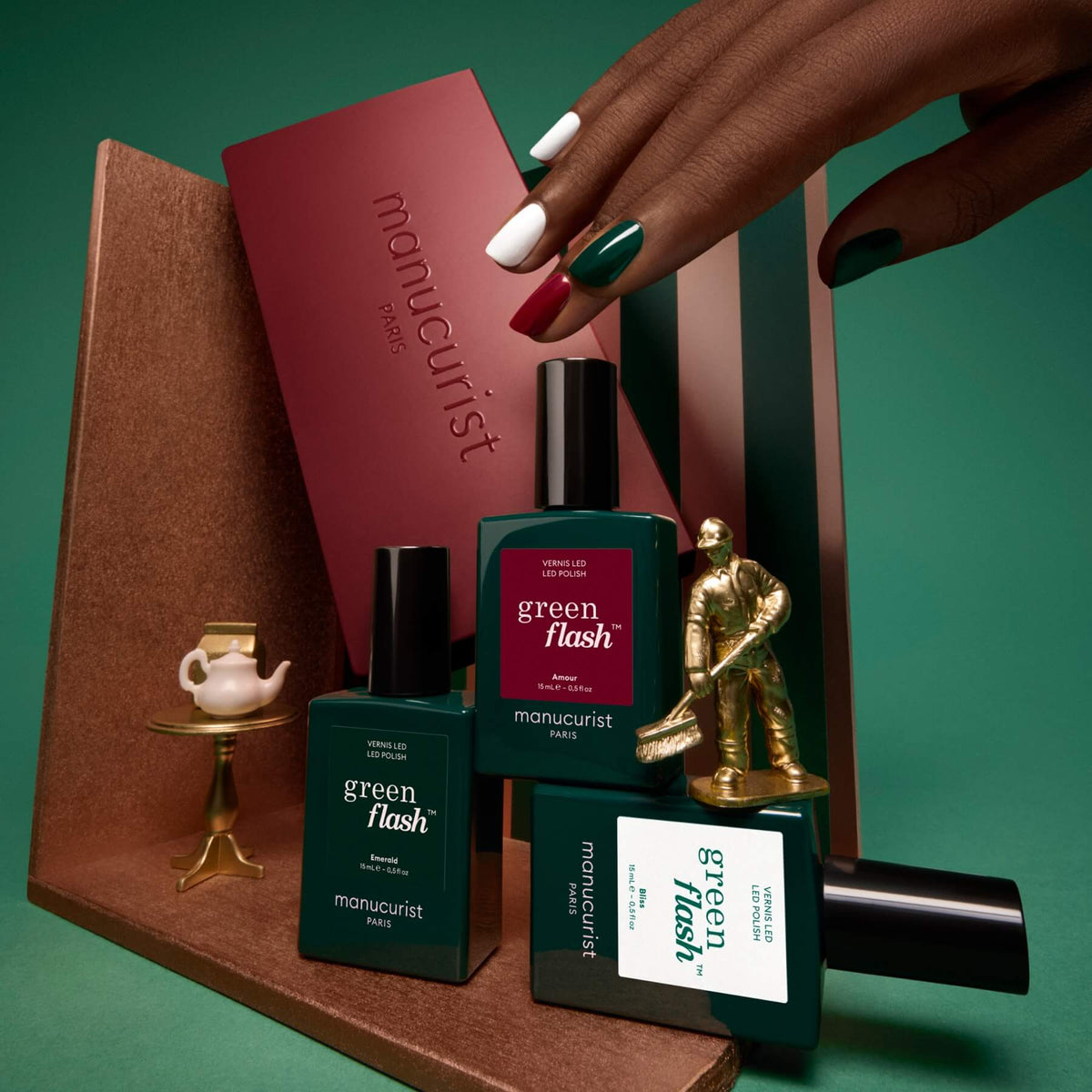 A hand with manicured nails in white, green, and burgundy touches a box of Manucurist’s Curated Trios. Three Green Flash nail polish bottles from the curated collection, along with decorative accents like a gold skater and a miniature teapot, are artfully displayed on a brown shelf against a green background—ideal manicure inspiration for anyone exploring the latest Green Flash color palettes by Manucurist.