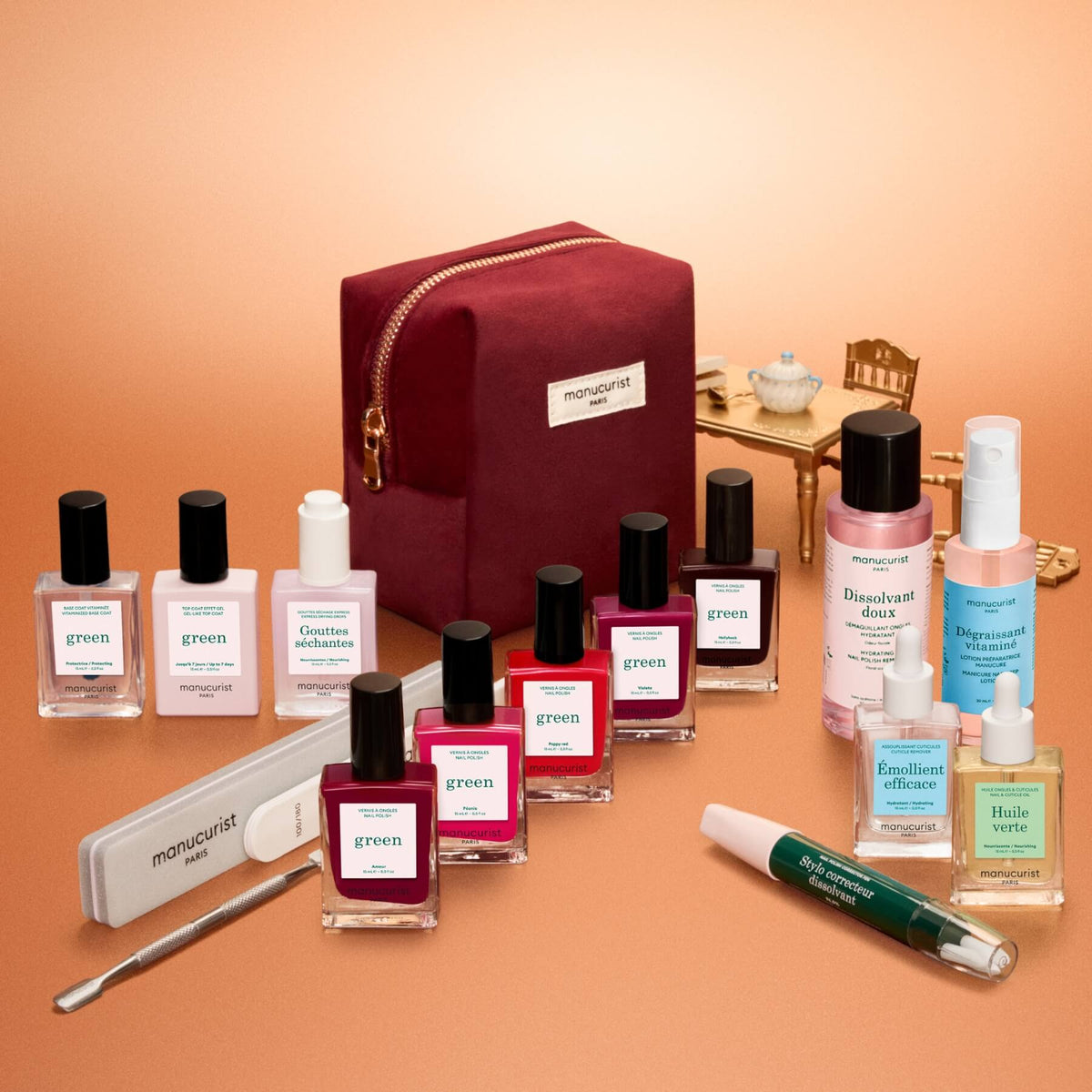A Manucurist Green™ Super Ultimate Kit Gift Set, featuring a maroon manicure bag surrounded by an array of naturally derived nail polishes, manicure tools, and care products, is displayed on a peach surface with miniature furniture in the background.