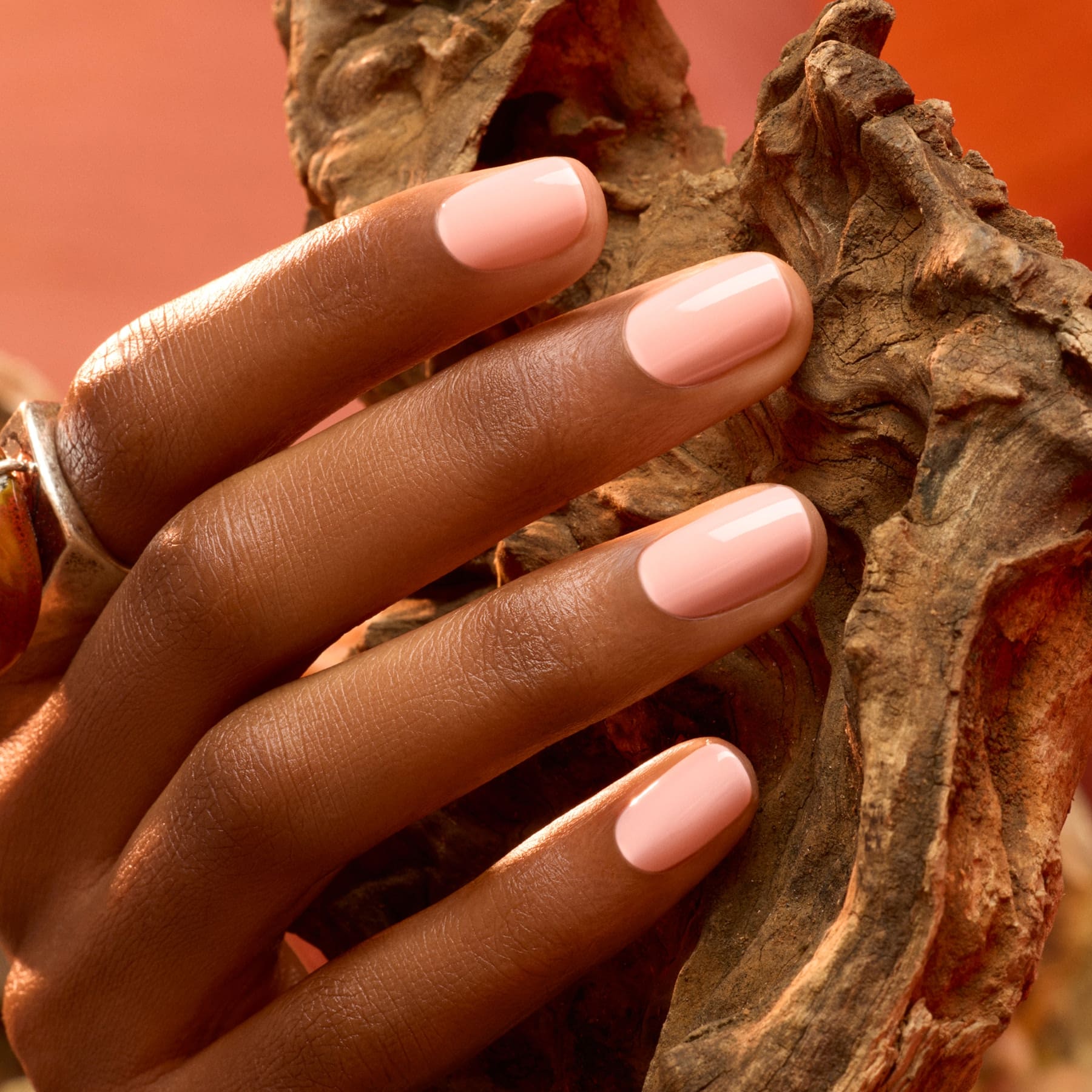 A hand with neatly manicured nails painted in Manucurist’s Milky Peach gel polish rests on a textured piece of wood; the background is warm and softly blurred.