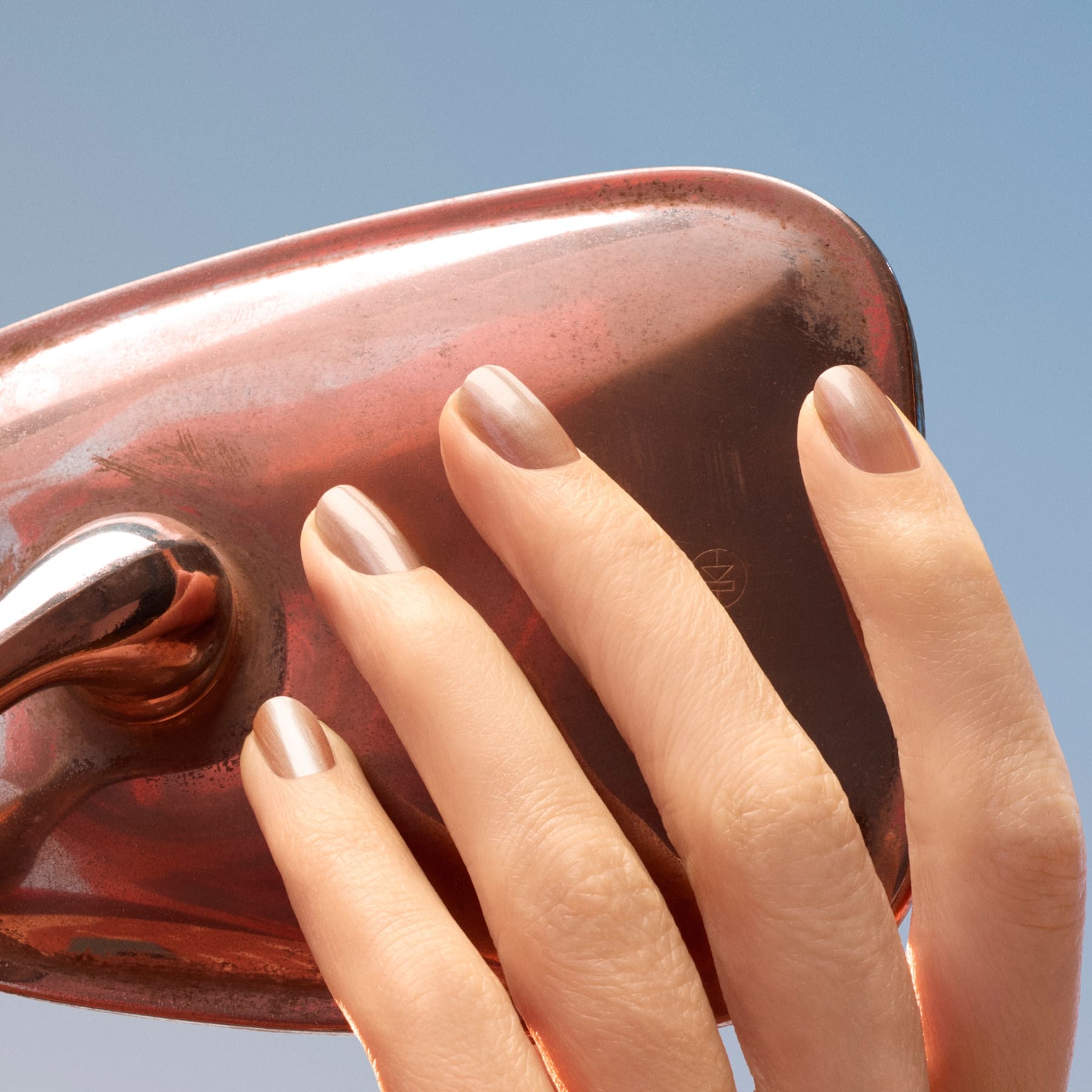 A hand with neatly manicured nails—painted in the shimmering latte brown shade “Sandstone” by Manucurist—touches a shiny, copper-colored object against a clear blue sky.