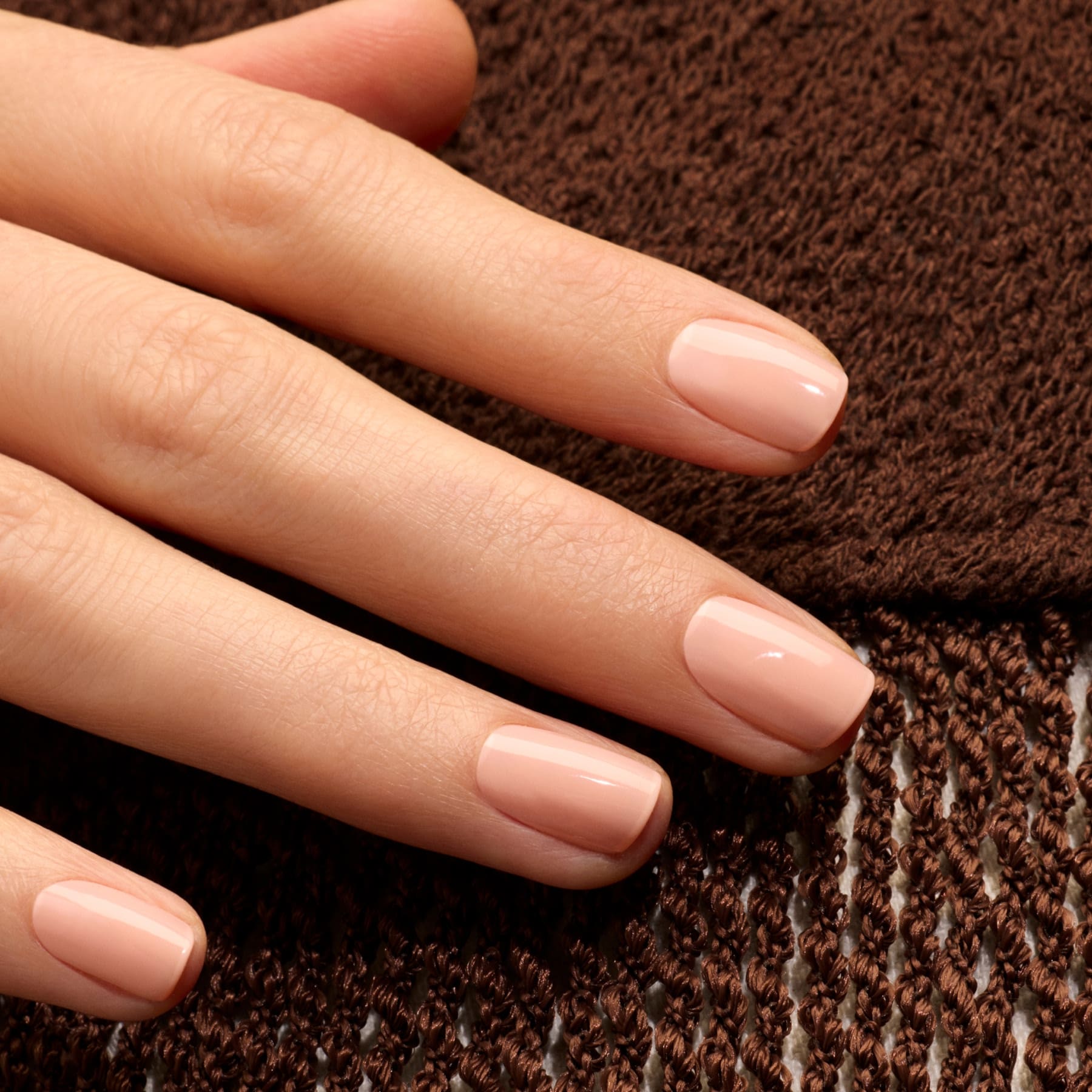 A close-up of a hand with neatly manicured nails painted in Manucurist’s Milky Peach gel polish, resting on brown textured fabric.