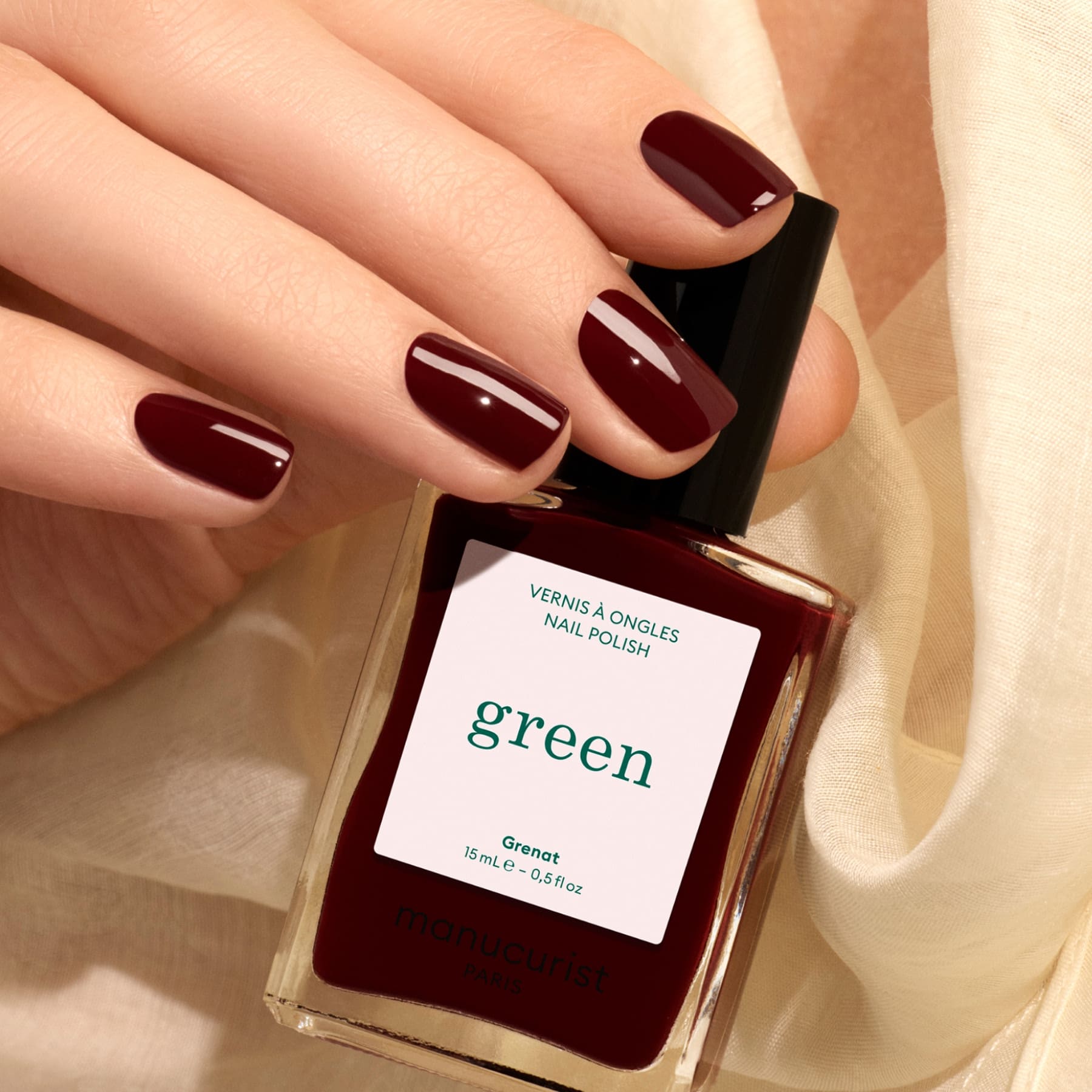 A hand with neatly manicured, maroon red nails holds a bottle of Manucurist Grenat nail polish. The background features a soft, cream-colored fabric.