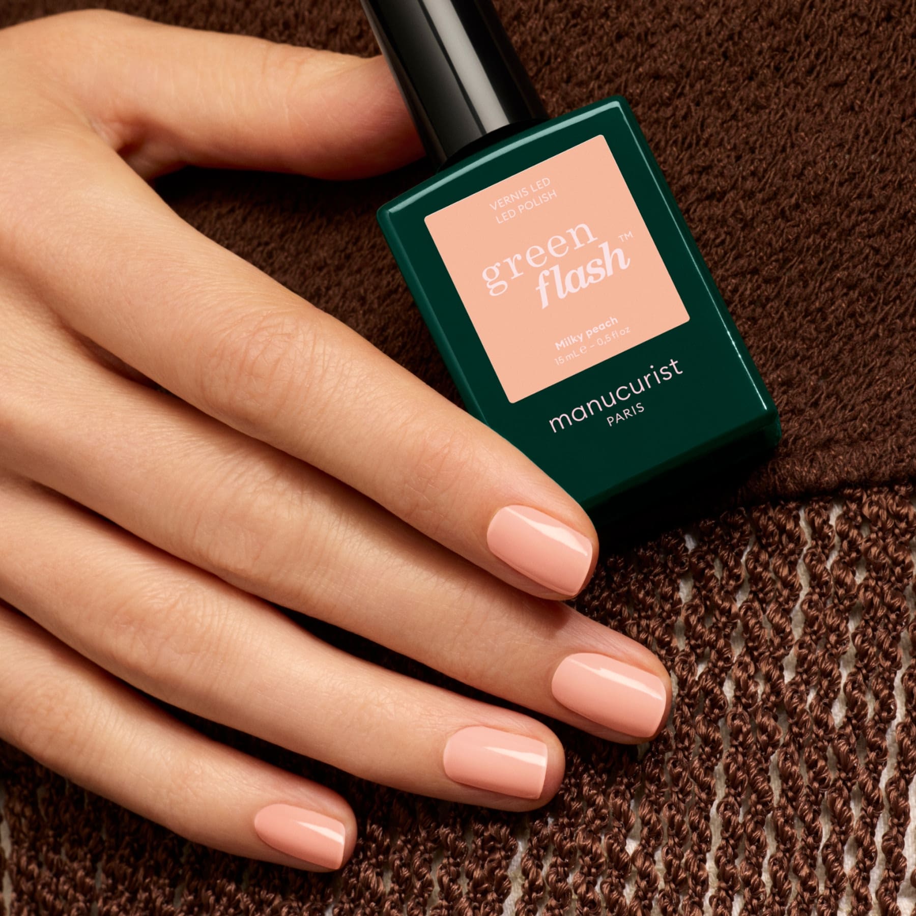 A hand with short, glossy, light pink nails—reminiscent of a milky peach gel polish—holds a bottle of Manucurist Milky Peach nail polish, resting on a brown textured fabric.
