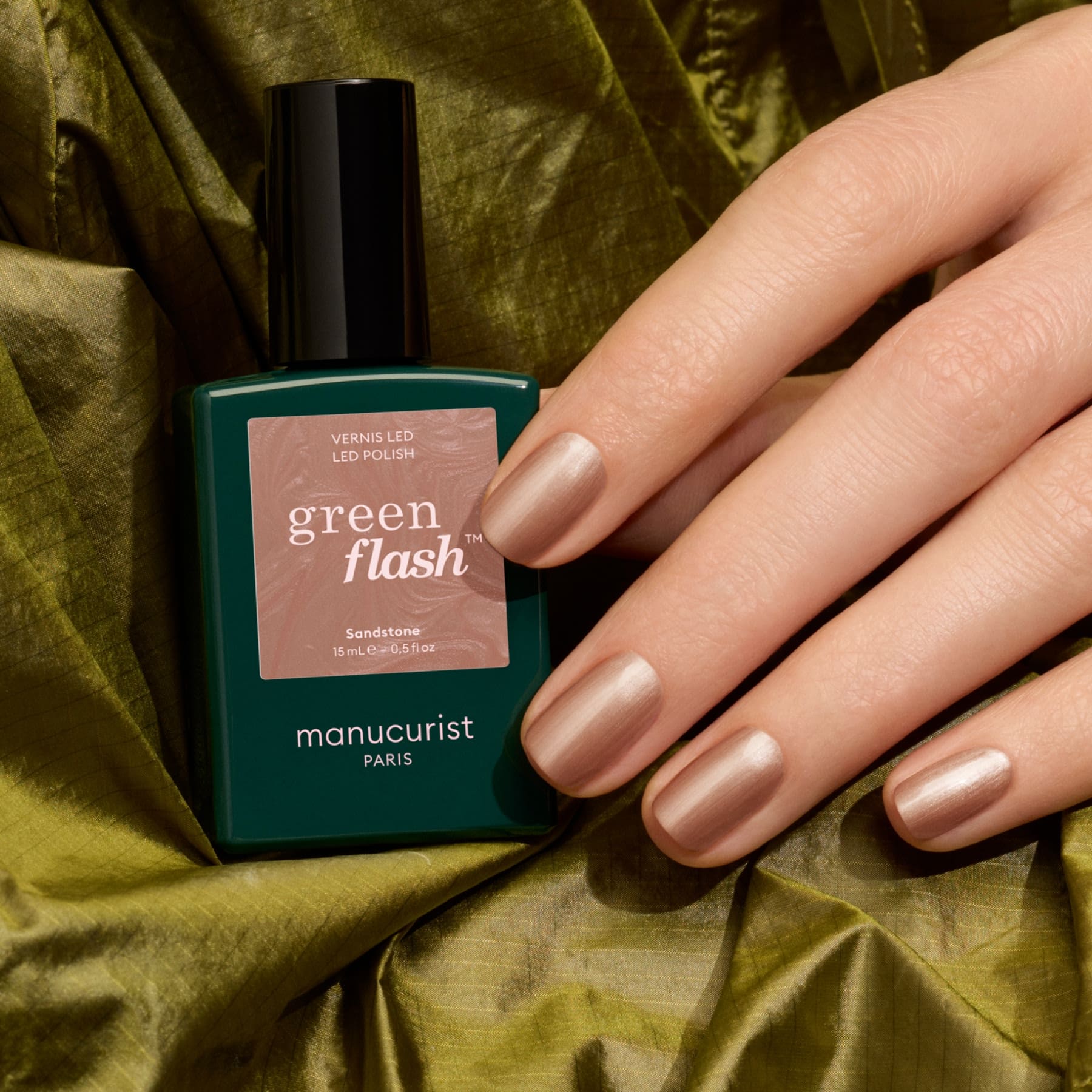 A hand with neatly manicured nails painted in a shimmering latte brown metallic nude shade holds a bottle of Manucurist Sandstone gel polish, resting on olive green fabric.