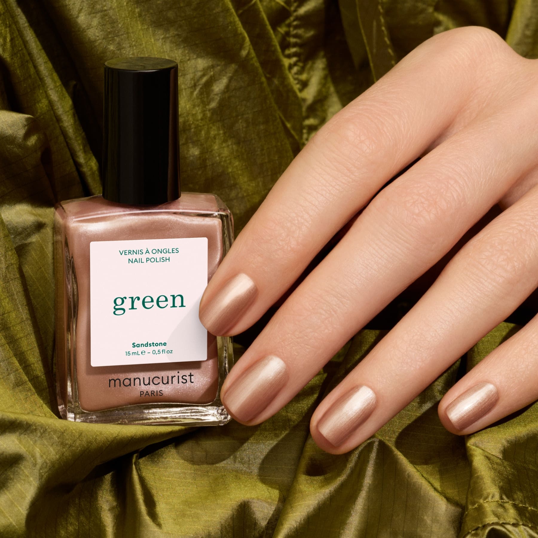 A hand with shimmery beige nail polish rests on green fabric beside a matching bottle labeled Manucurist, shade Sandstone. The soft glow of the Sandstone polish evokes the elegance of pearly brown nail colors, adding a refined touch to the look.