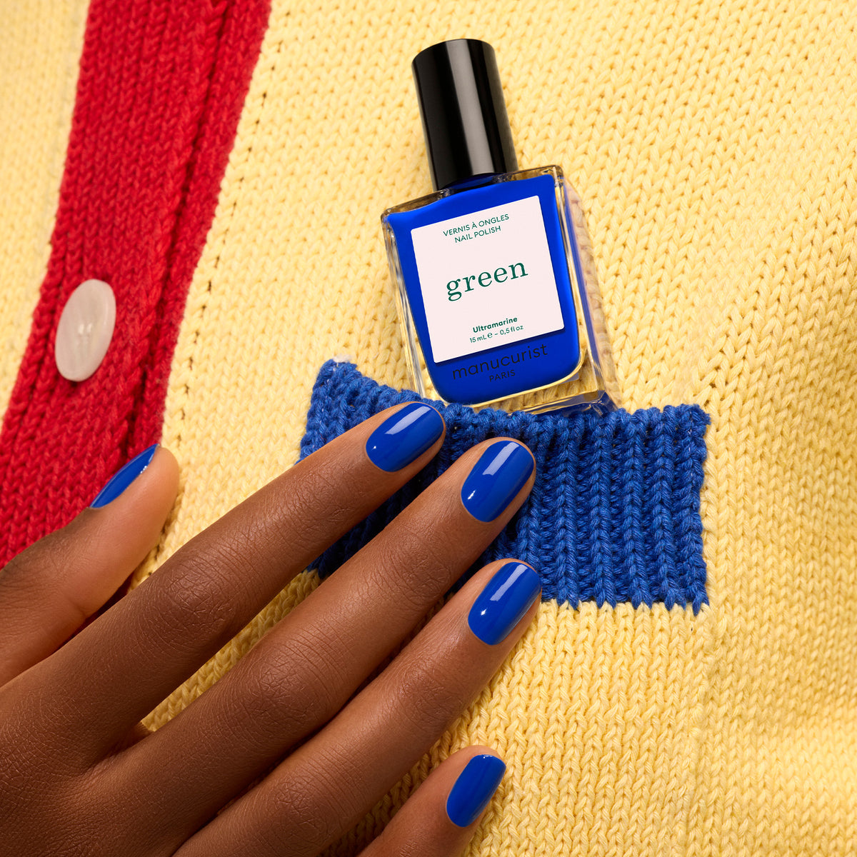 A hand with Manucurist Ultramarine electric blue nail polish holds the matching bottle in the pocket of a yellow knit sweater with a red button and blue trim.