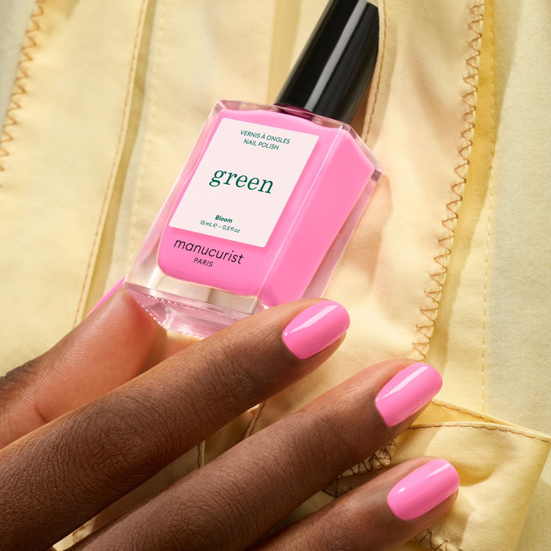 A hand with glossy neon pink nail polish, featuring blue undertones, holds a matching pink bottle of Manucurist Bloom nail polish against a light yellow fabric background.
