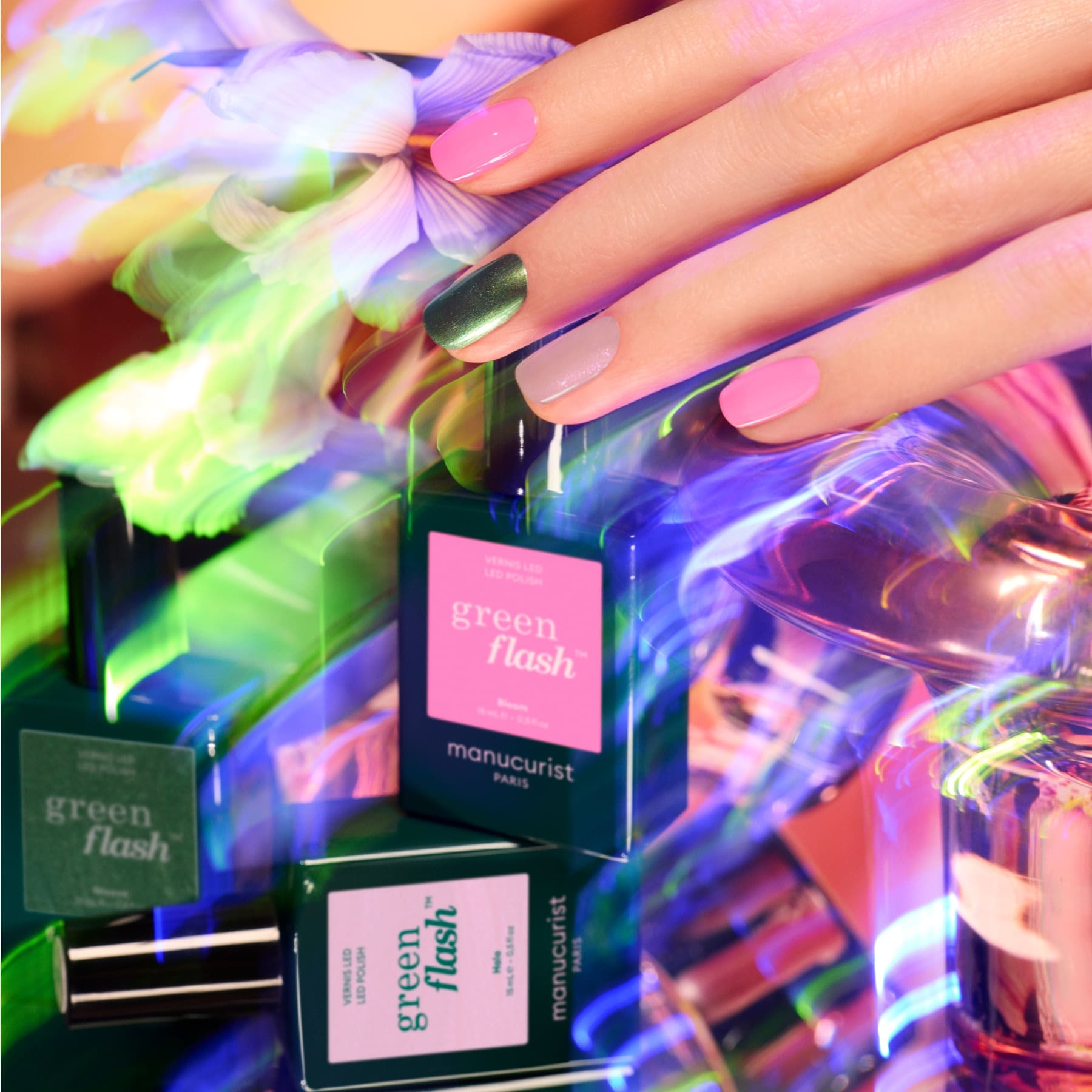 A hand with pink and green nails rests above colorful Manucurist Curated Trios bottles, surrounded by vibrant light reflections—perfect inspiration for new Curated Trios color palettes and manicure trends.