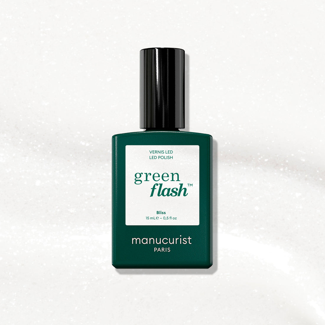 Un flacon de vernis gel Bliss de Manucurist, avec une étiquette vert foncé et un bouchon noir, est présenté sur une surface blanche irisée aux reflets subtilement scintillants.