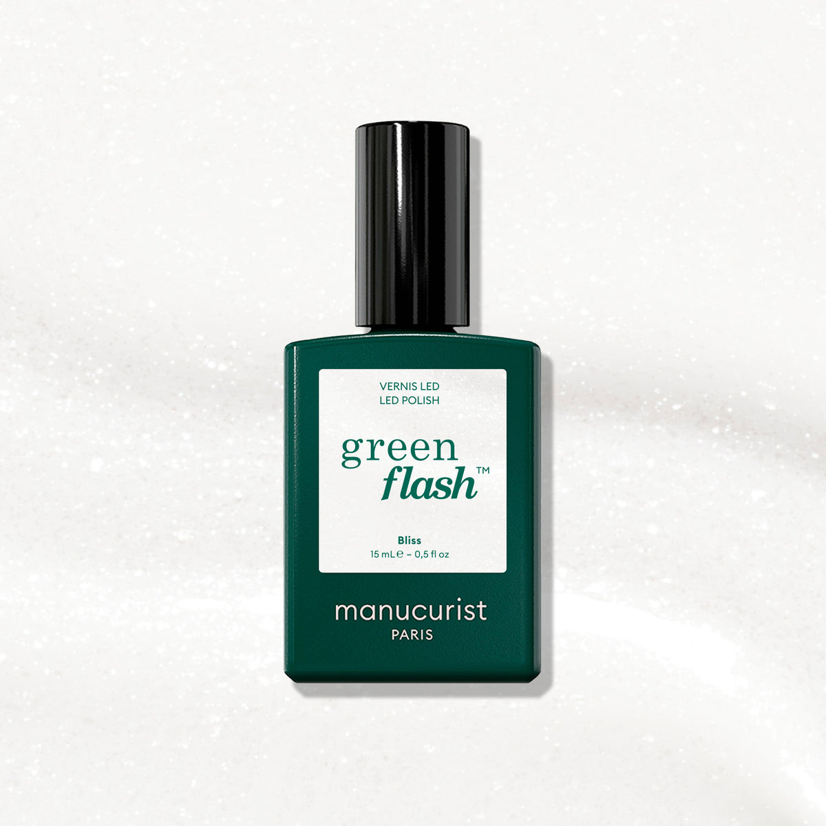 A bottle of Manucurist Bliss gel polish, featuring a dark green label and black cap, is displayed on an iridescent white, subtly sparkling surface.