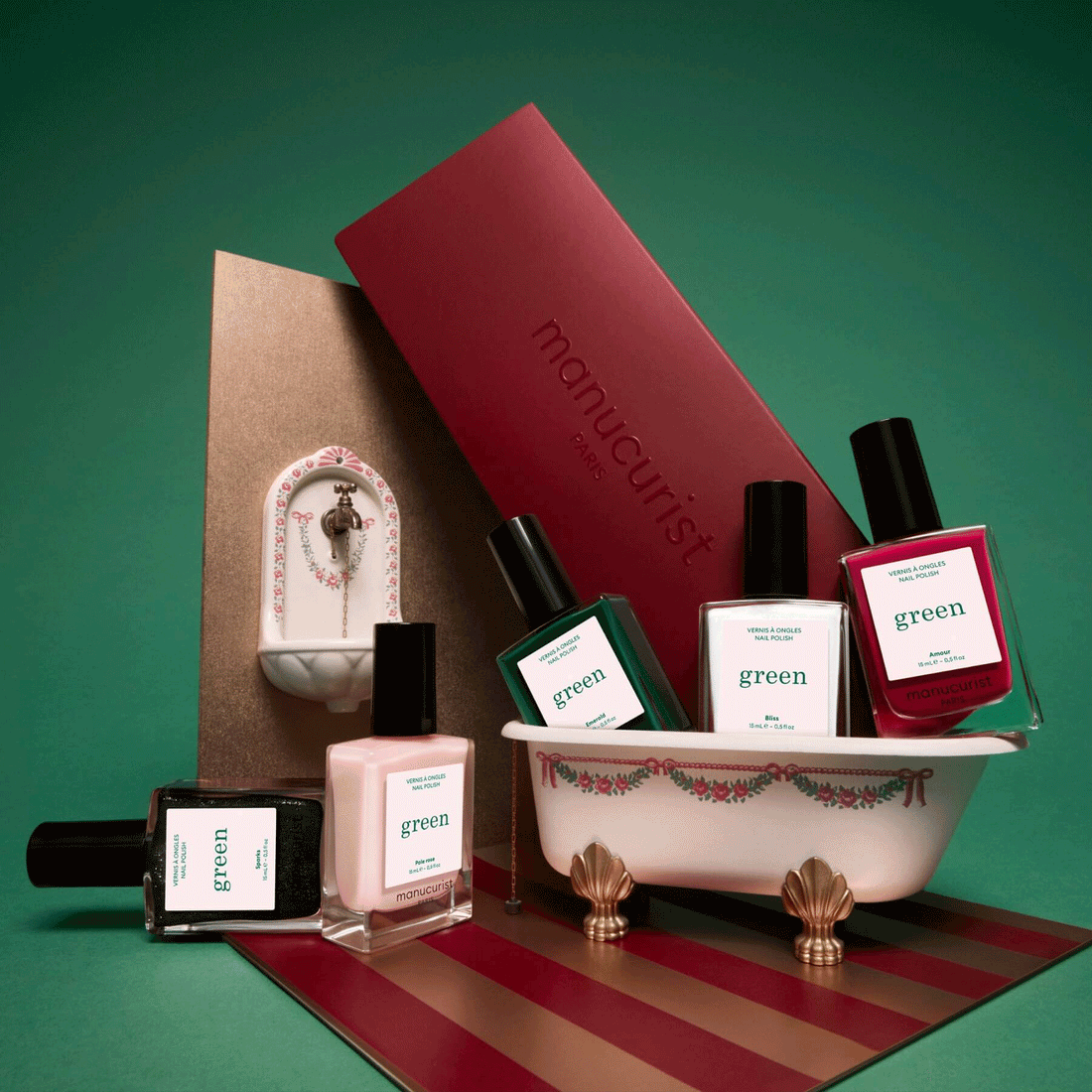 Five bottles from the Manucurist Customizable 5-pack, featuring a striking green polish, are artfully arranged around a small decorative bathtub. In the background, a red manicurist box and a miniature sink complete the scene—all set against a vibrant green backdrop. This playful arrangement highlights the fun of creating your own personalized rainbow nail set with Manucurist.