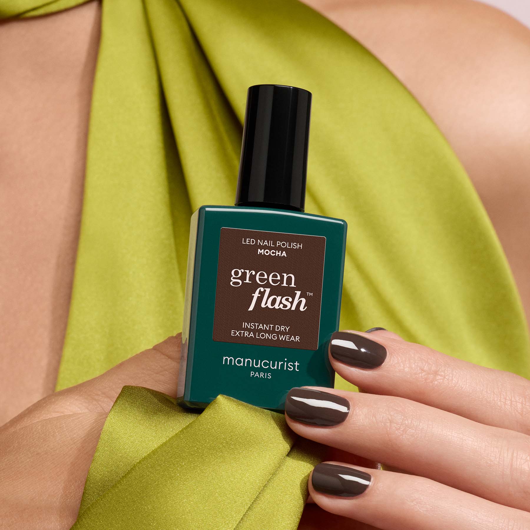 A hand with glossy dark brown Manucurist Mocha nail polish holds a bottle of the same shade, showcasing cool undertones. The person is wearing a lime green garment draped over their shoulder.