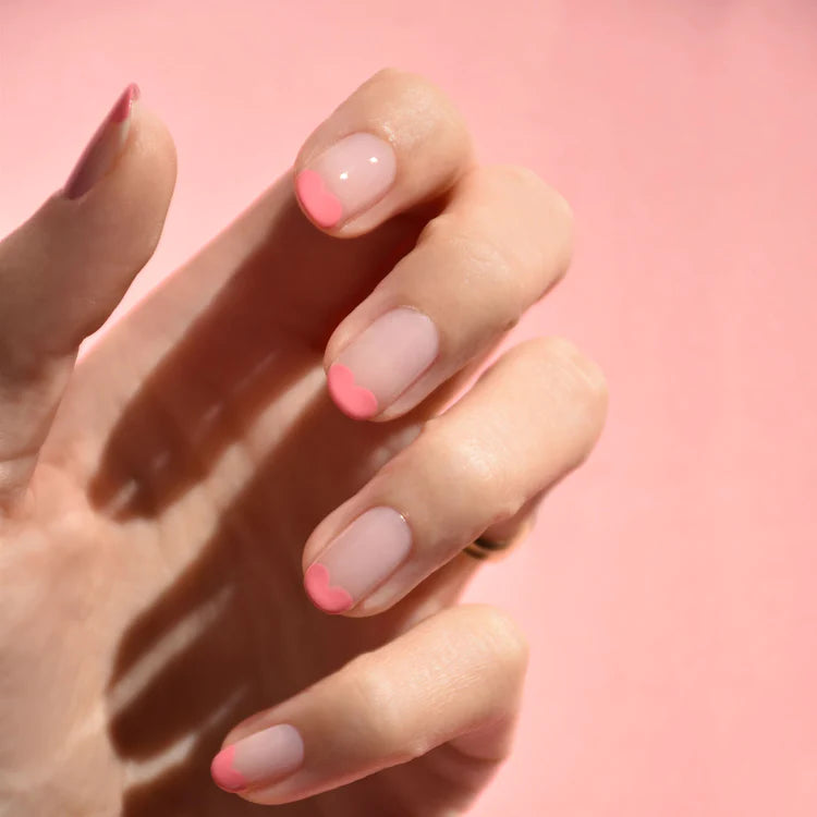 A hand with manicured nails features a light pink base and heart-shaped pink tips, set against a soft pink background.