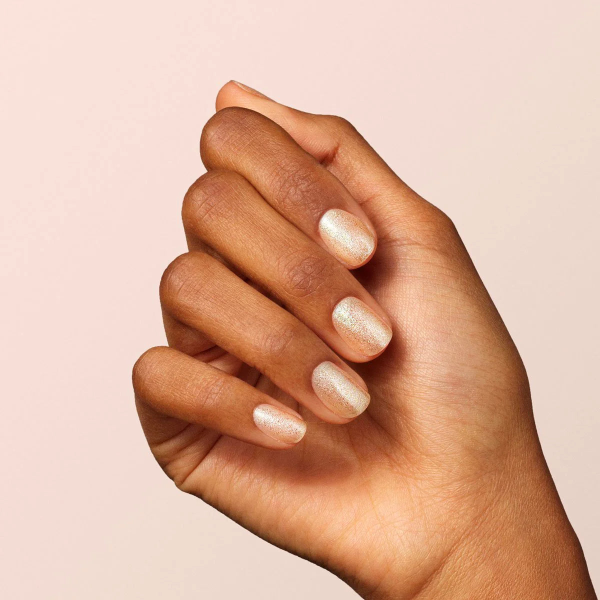 A hand with medium brown skin shows neatly manicured nails painted with a shimmery, nude-colored polish, set against a light beige background.