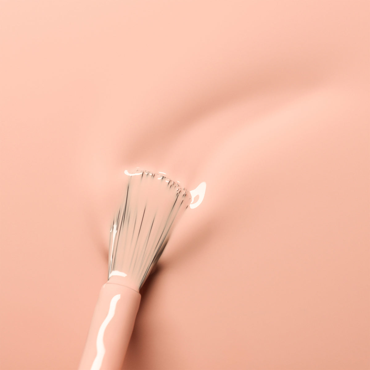 Un pinceau de maquillage aux poils blancs tourbillonne sur une surface lisse rose pêche, révélant la teinte Pink Sand douce et brillante de Manucurist — un look épuré et minimaliste aux subtils sous-tons orangés.