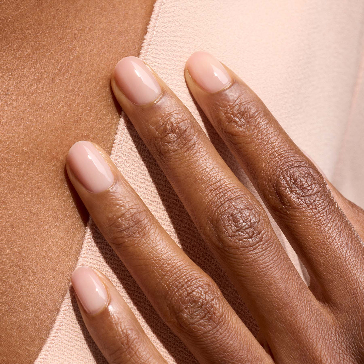 A close-up of a hand with short, oval-shaped nails polished in Manucurist’s Pale Rose, a sheer pastel pink nail polish, shows elegantly manicured, natural-looking nails resting on light beige fabric and skin for an understated look.