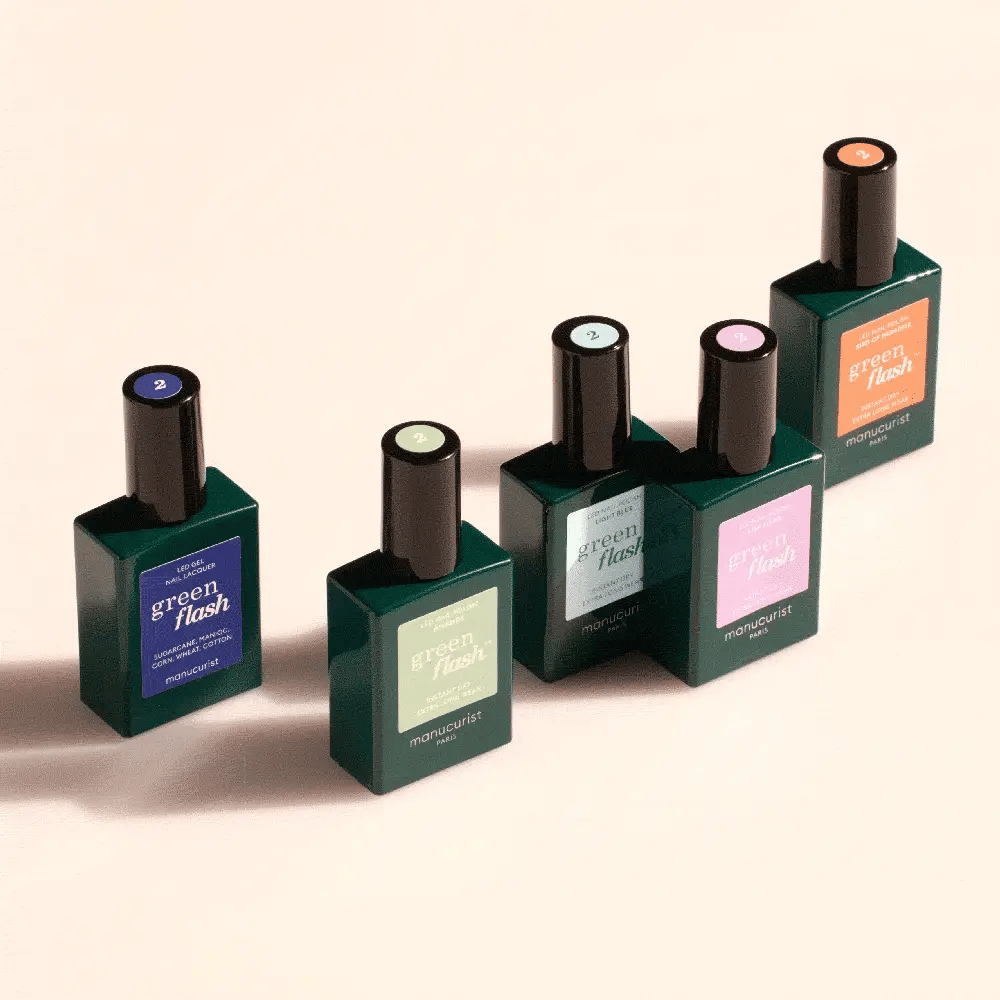 Five Montmartre Green Flash nail polish bottles in dark green, each with a different colored label and cap, are arranged upright on a light beige surface, casting soft shadows.