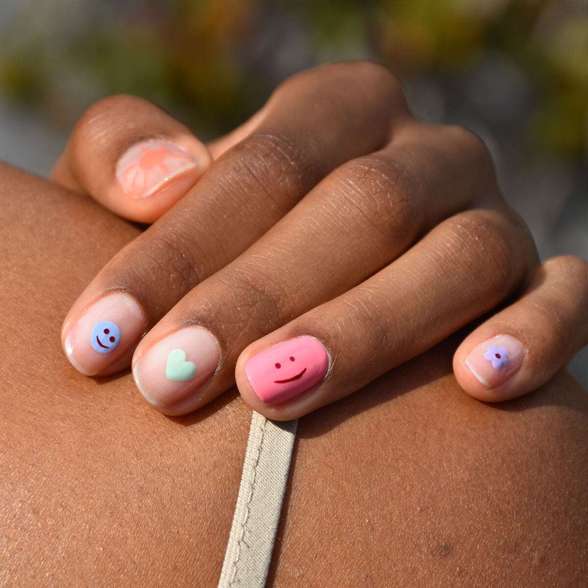A close-up of a hand with nude nails featuring playful nail art: blue and pink smiley faces, a white heart, and a small purple flower. The hand rests on a shoulder with a beige strap visible.