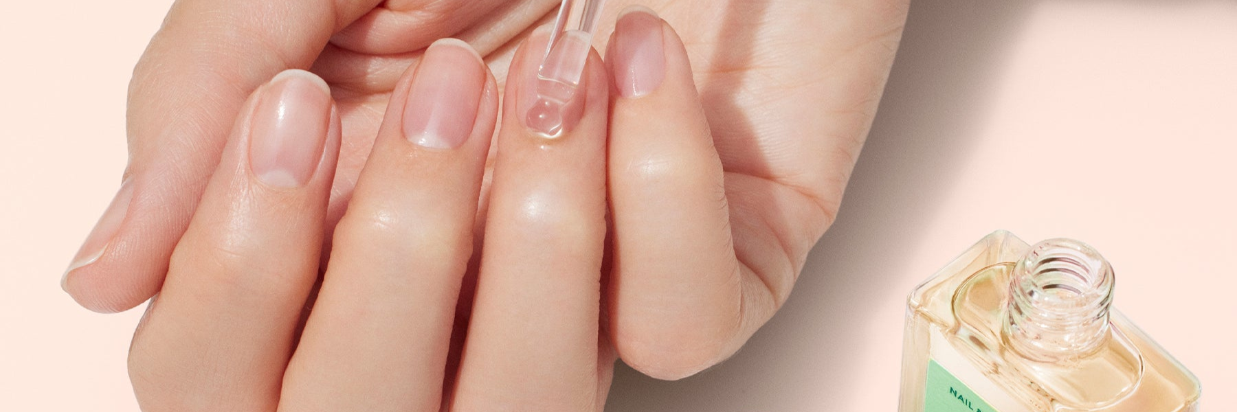 A hand applies cuticle oil to fingernails using a dropper, with an open bottle of oil nearby on a light background.