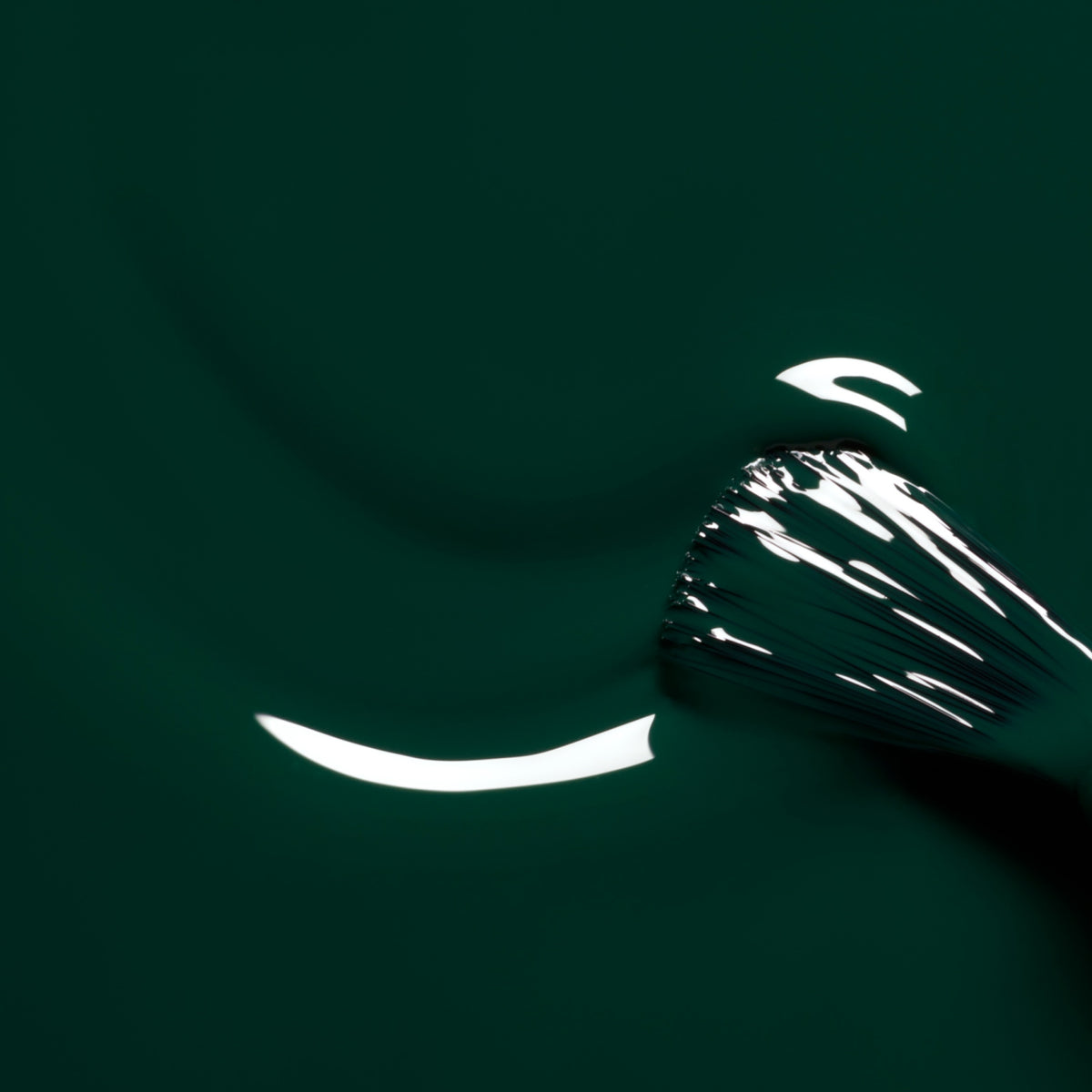 A close-up of a makeup brush swirling through glossy Manucurist “Emerald” nail polish, its deep forest green shade creating smooth, reflective curves and luminous highlights on the surface.