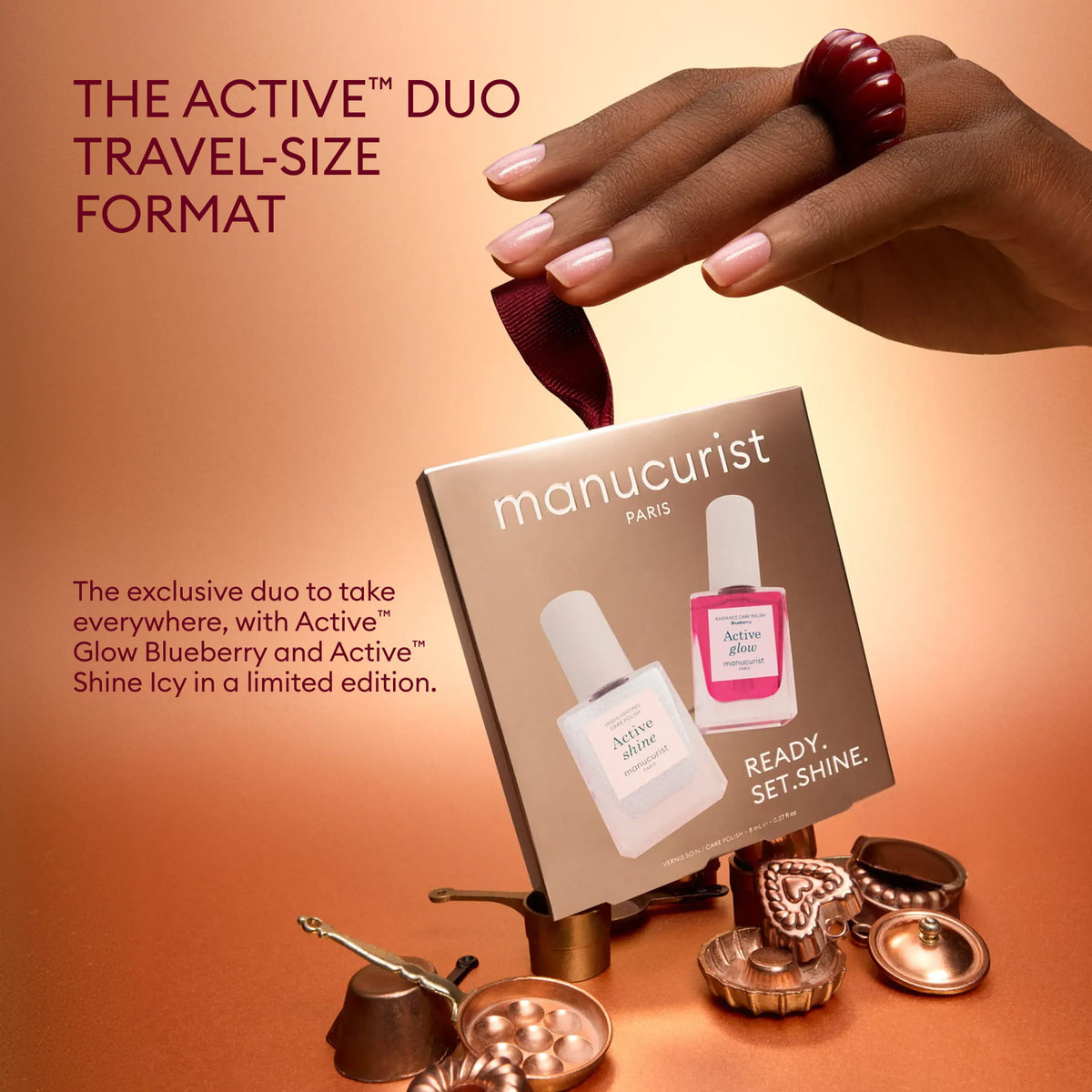 Une main manucurée tient la boîte du duo format voyage Active™ de Manucurist, un coffret exclusif en édition limitée pour les fêtes, parfait pour les passionnés de soin des ongles. À l’arrière-plan, de petites coupes cuivrées et un ruban donnent une touche festive à ce cadeau Manucurist.