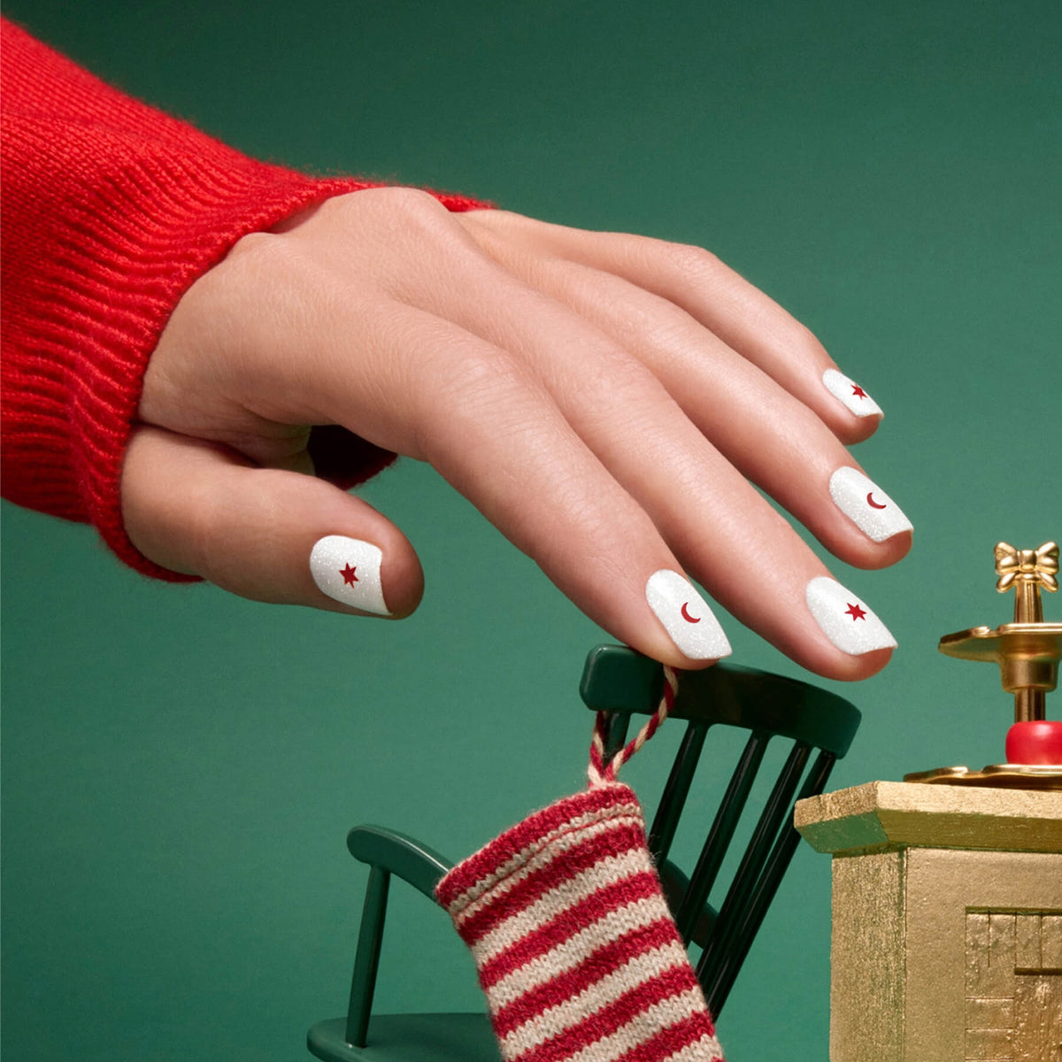 A hand adorned with Manucurist Stickers Nail Art Holidays—white polish decorated with red stars and crescent moons—peeks out from a red sweater, hovering above a small green chair and a gold ornament wearing a striped red and white stocking, offering perfect festive manicure inspiration.