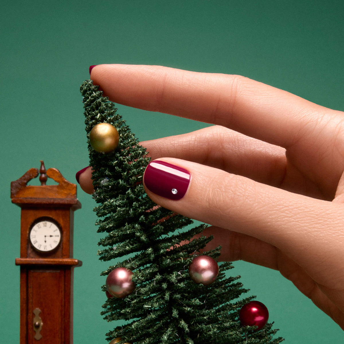 A hand with a flawless burgundy manicure, adorned with Manucurist’s Rhinestones Nail Art Holidays, decorates a small Christmas tree with ornaments—infusing festive charm inspired by holiday looks. A miniature grandfather clock stands beside the tree, all set against a green background.