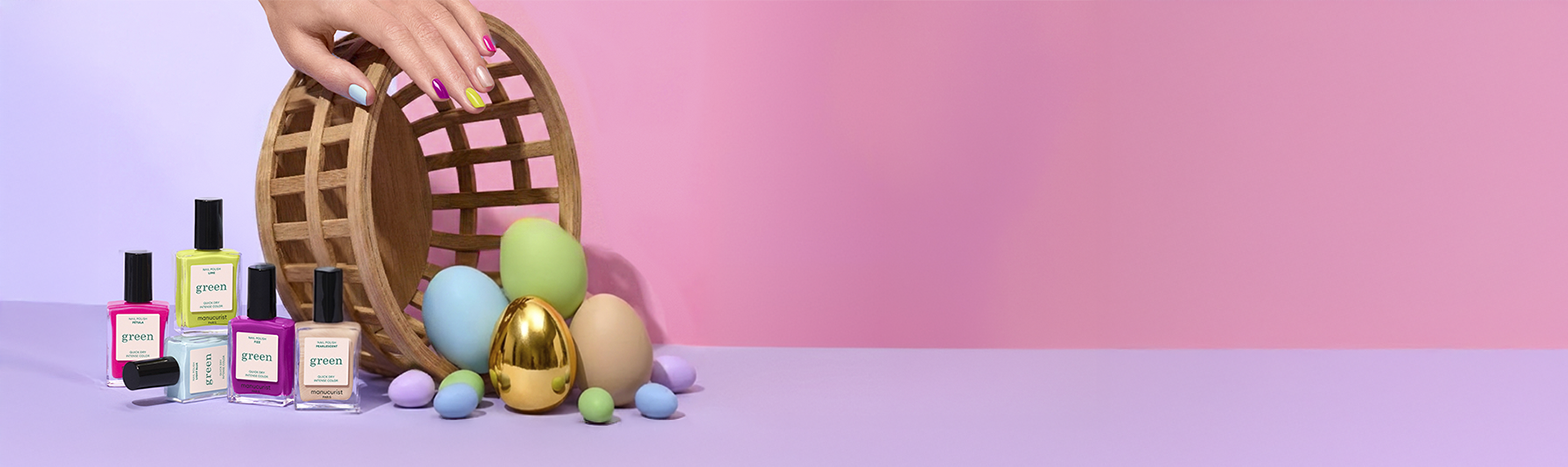 A hand with pastel-painted nails tips over a wicker basket, spilling out colorful eggs, a gold egg, and several bottles of nail polish on a purple surface against a pink and purple gradient background.
