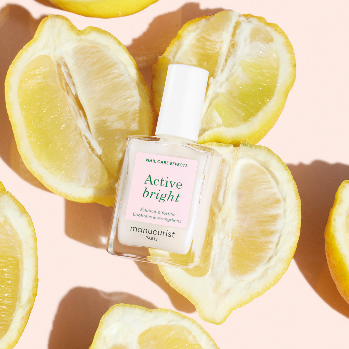 Un flacon de vernis soin Active™ Bright Manucurist repose sur un fond pêche, entouré de citrons coupés en deux disposés en cercle ouvert. La lumière du soleil filtre à travers, projetant de douces ombres des citrons et du flacon.
