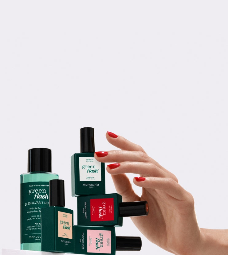 A hand with red nail polish reaches towards a stack of green bottles and nail polish, labeled green flash by Manucurist, against a light gray background.