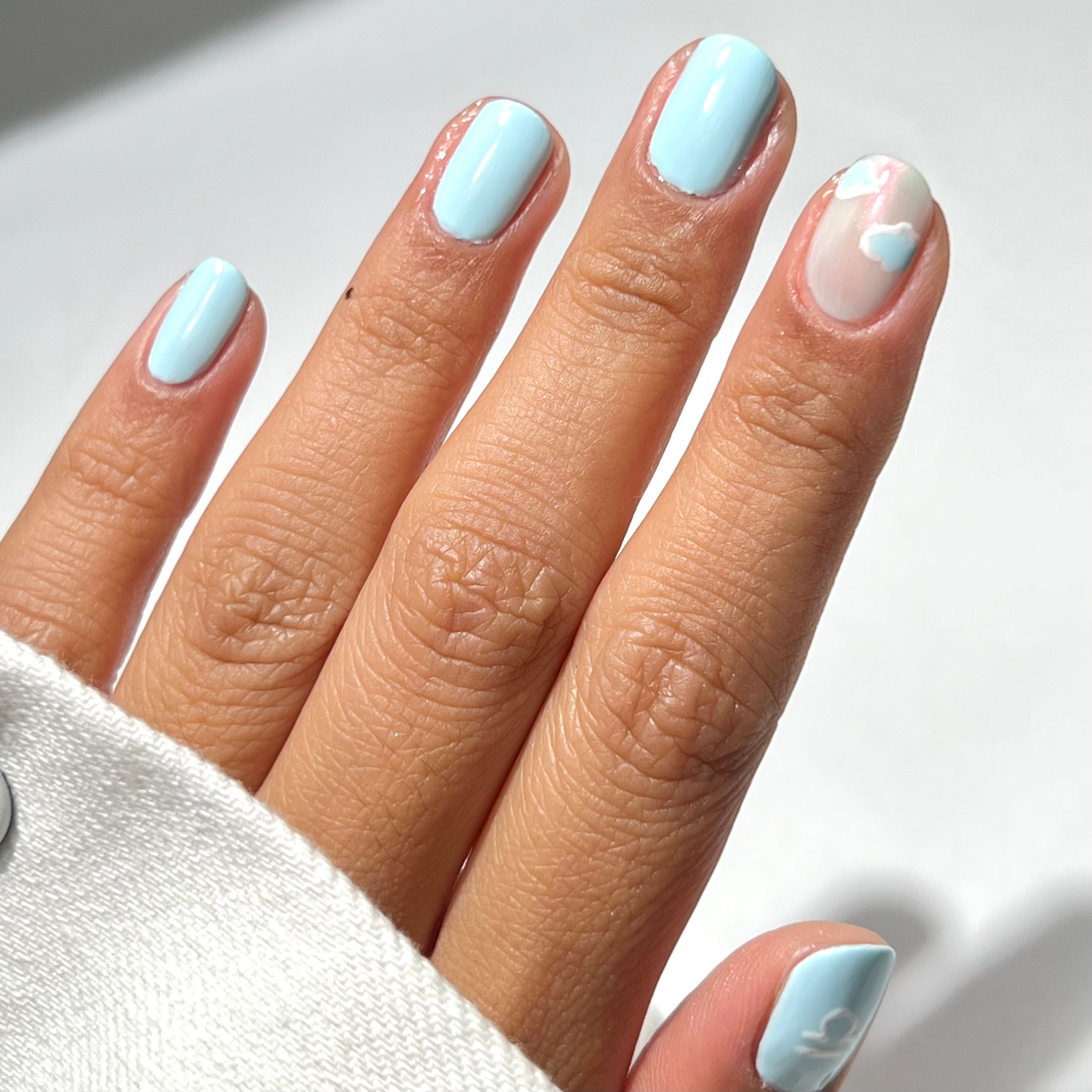 A hand with light blue nail polish; the ring finger has a white base with pastel blue and pink cloud-like designs. The person wears a white sleeve.