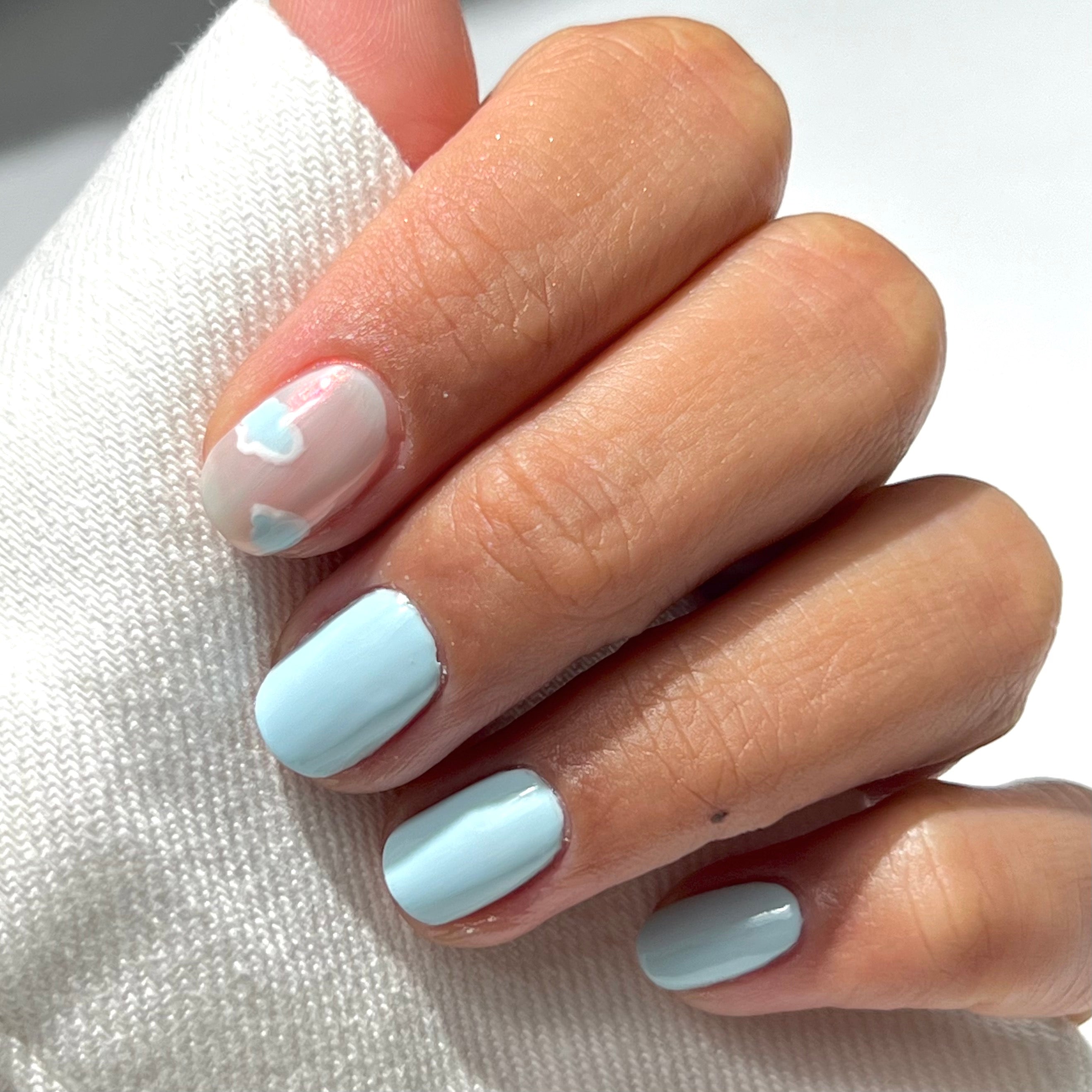 A hand with light blue nail polish on three fingers, while the index finger has a clear base with two small white heart designs. The hand is resting on white fabric.