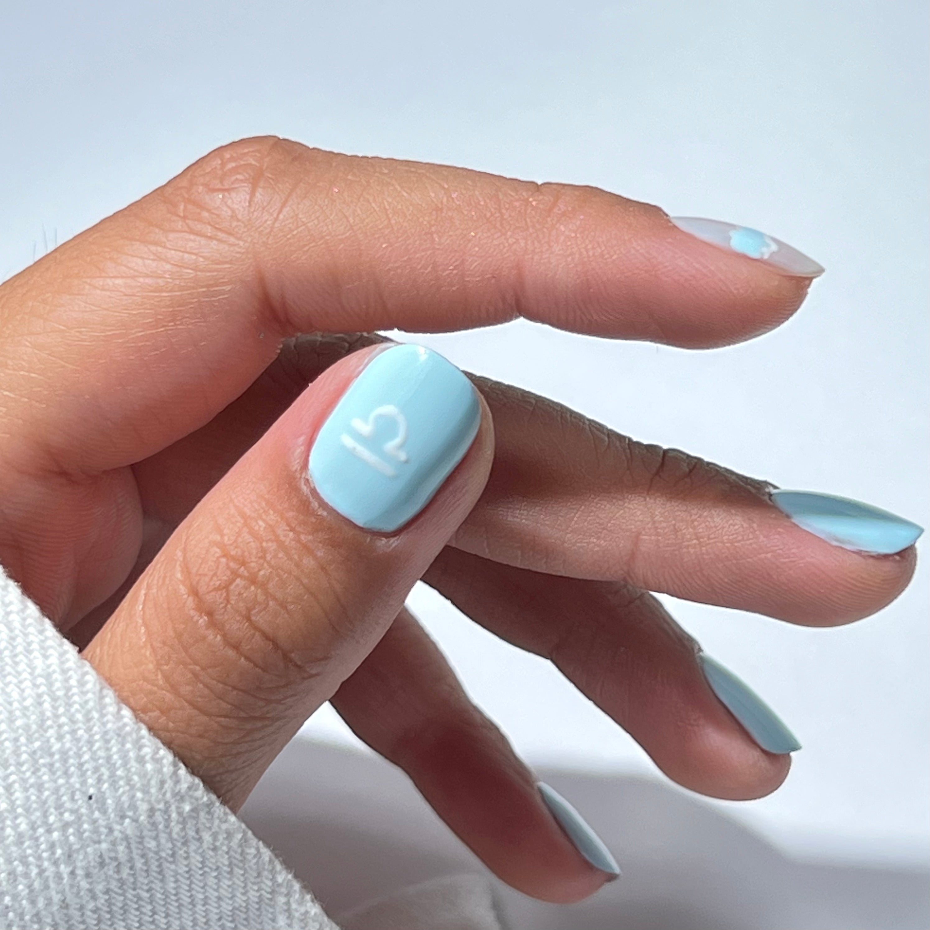 A hand with light blue nail polish, featuring a white Libra zodiac symbol on the ring fingernail. The background is plain and the person is wearing a white sleeve.
