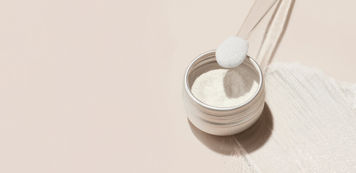A small jar of white powder with a clear scoop resting on top, some powder spilled beside it on a light beige surface.