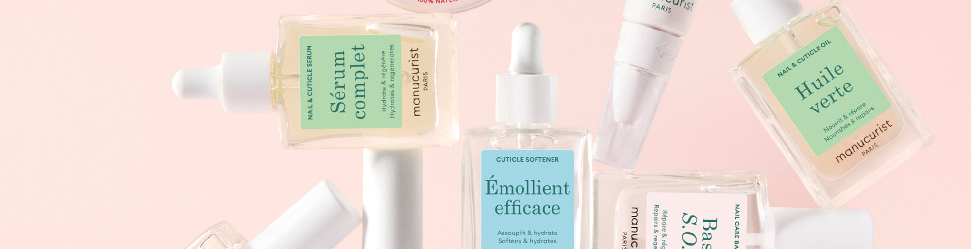 Various clear and light-colored nail care bottles and droppers with pastel green and blue labels are arranged artistically against a soft pink background.