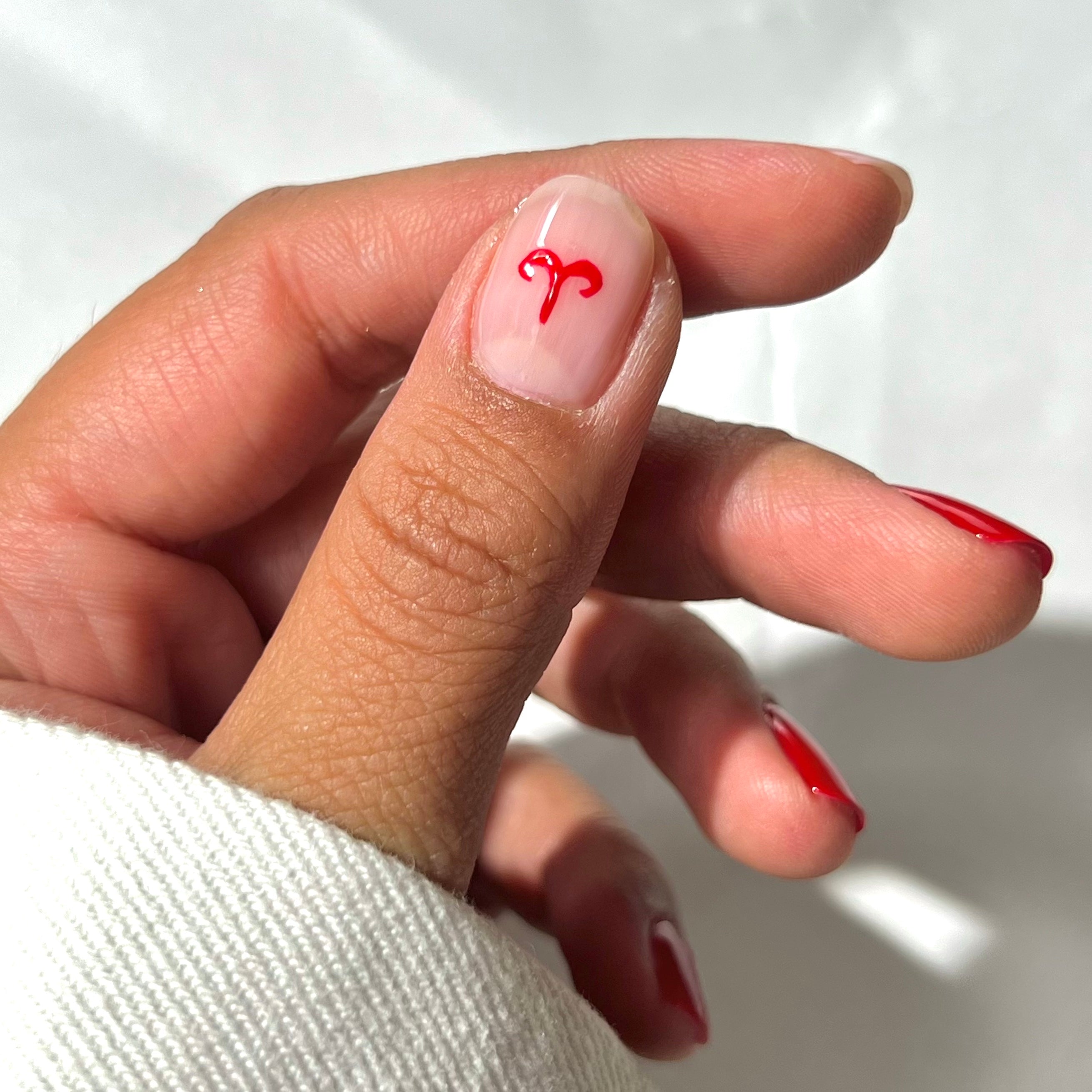 A hand with short, rounded nails painted red, except for the thumbnail, which is nude with a small red Aries zodiac symbol near the cuticle. The hand is partly wrapped in a white sleeve.