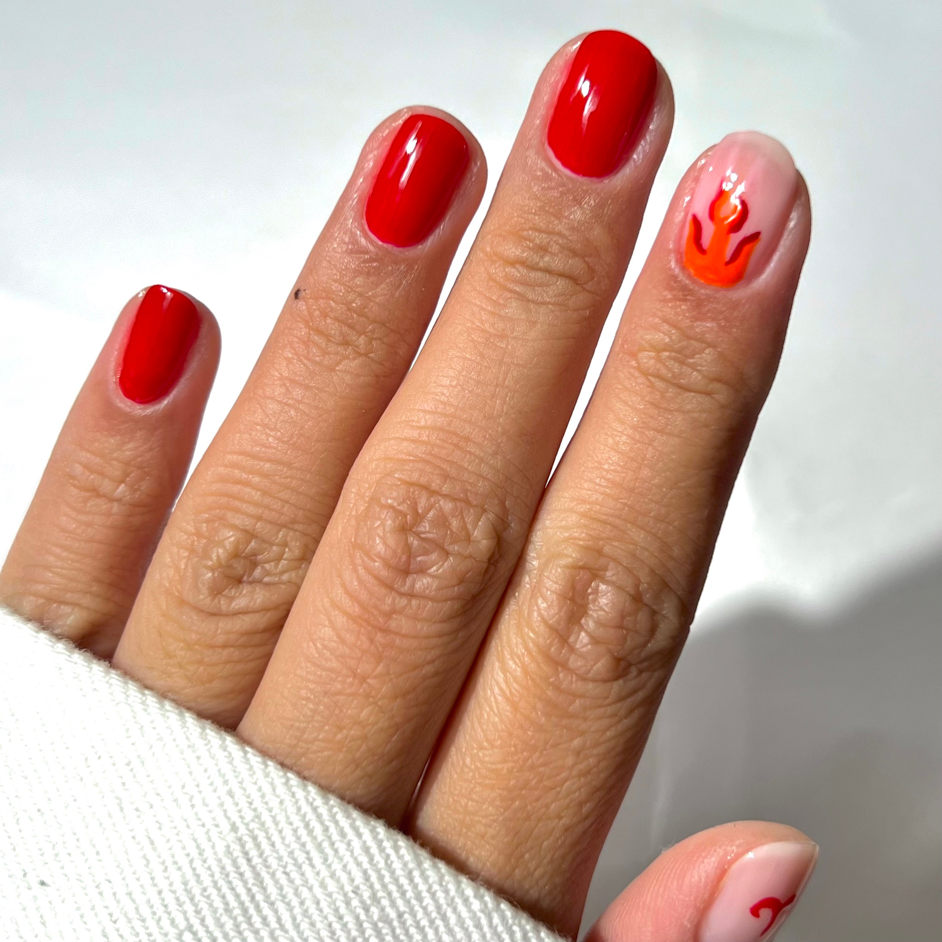 A hand with short nails painted red; the ring fingernail is light pink with an orange and red flame design. The hand is partially covered by a white sleeve.