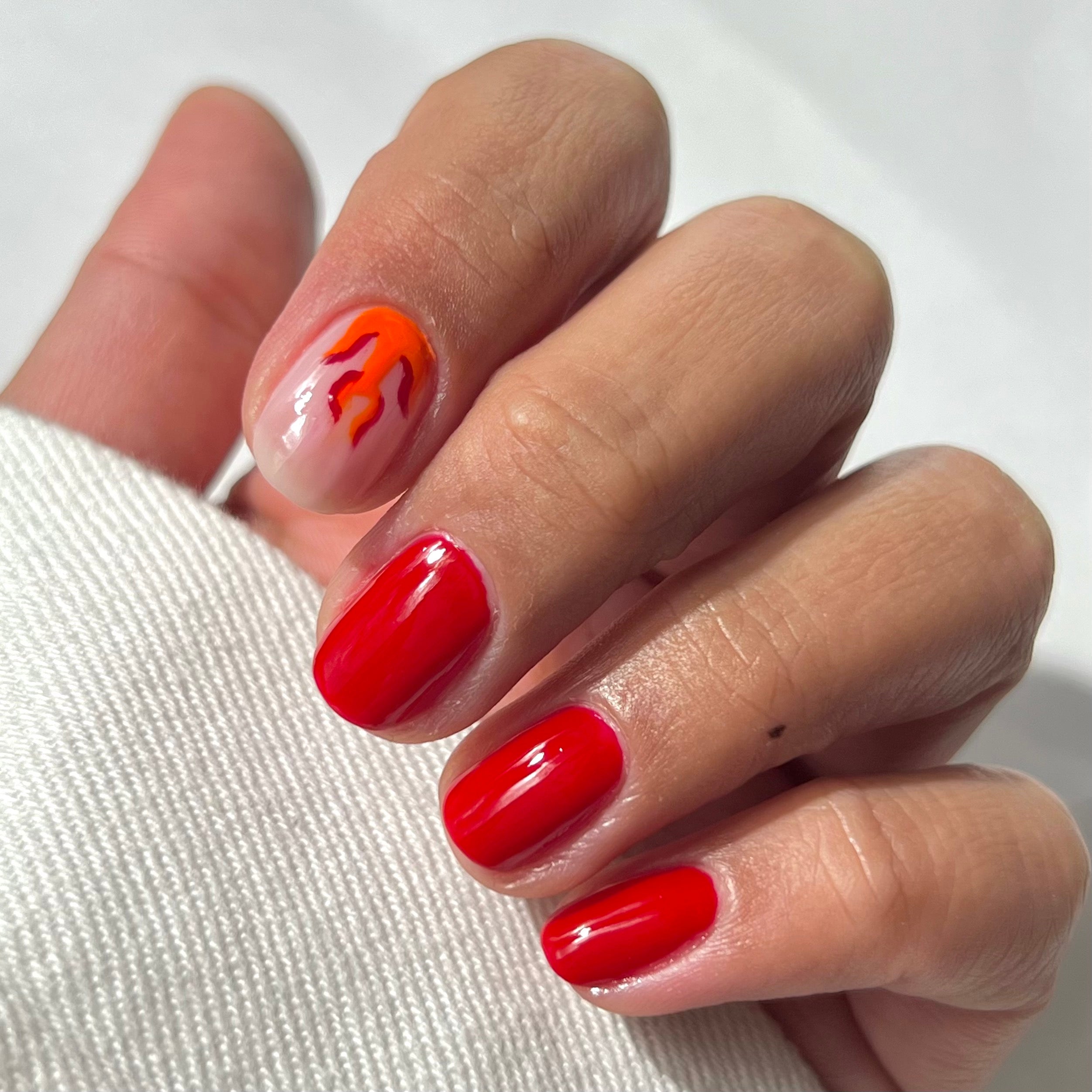 A hand with neatly manicured nails; four nails are painted glossy red, while the index finger has a clear base with an orange and red flame design. The hand rests on a white fabric.