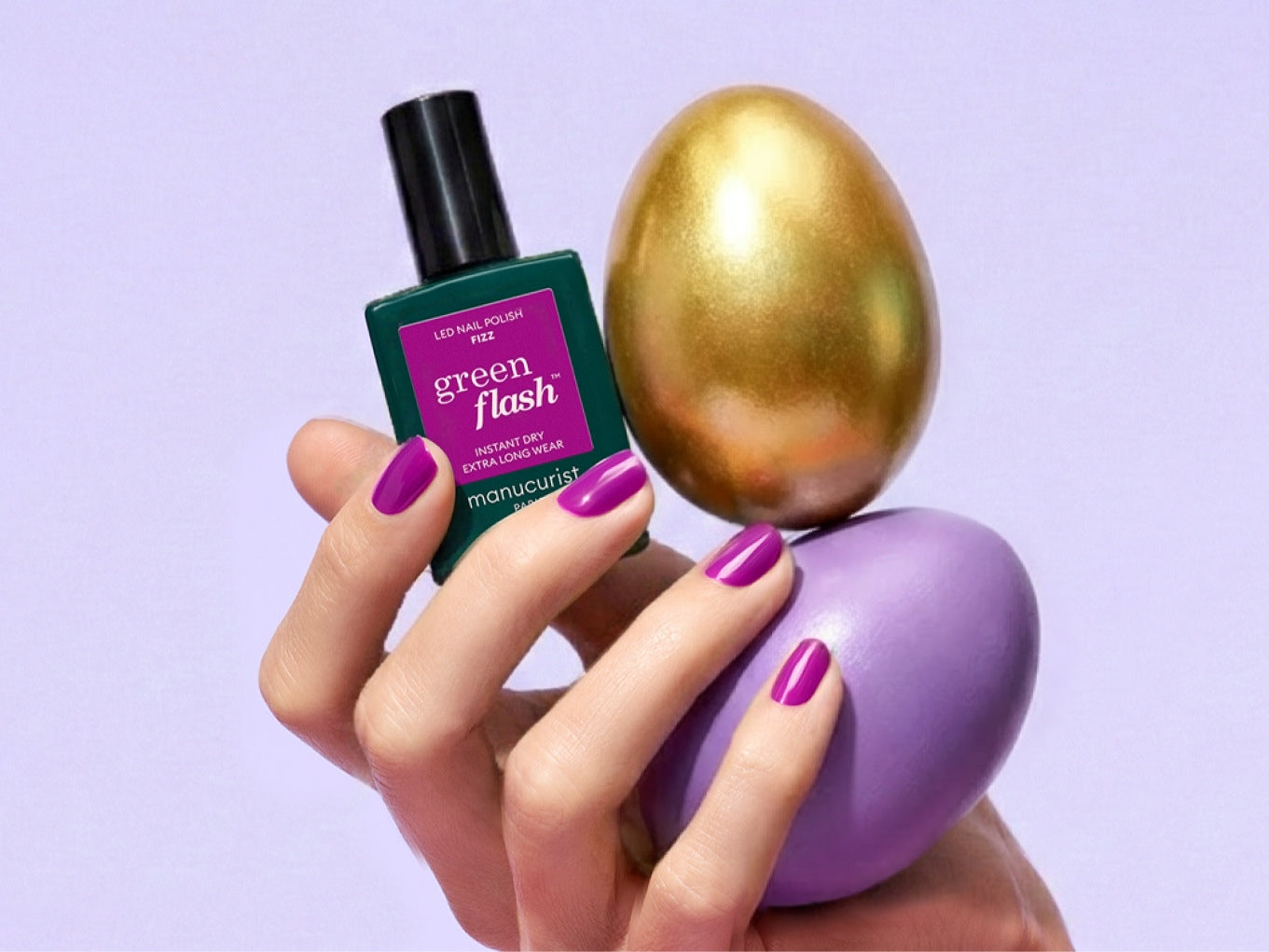 A hand with purple-painted nails holds a bottle of green flash nail polish and balances a gold egg and a purple egg against a pastel background.