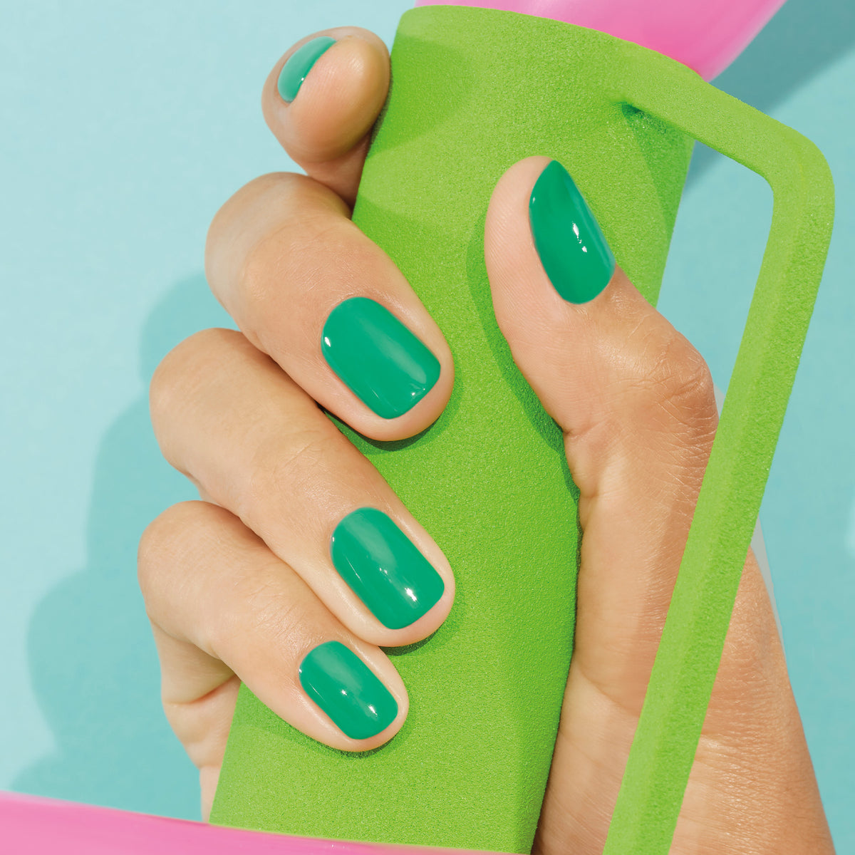 Une main aux ongles soigneusement manucurés, peints avec Brazil de Manucurist, une teinte vert électrique, et finis avec Top Coat, tient une poignée en mousse verte sur un fond bleu clair.